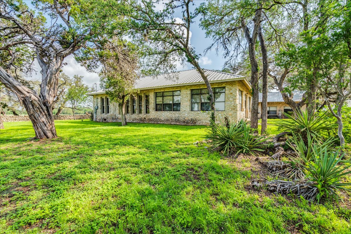 6400 Cypress Road Kyle, TX 78640 - Photo 6 of 33 Stone exterior residence featuring a metal roof, multiple windows, and a surrounding landscape with mature trees and varied plantings