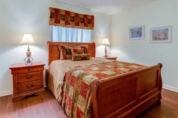 a bedroom with a bed and a lamp on dresser with wooden floor
