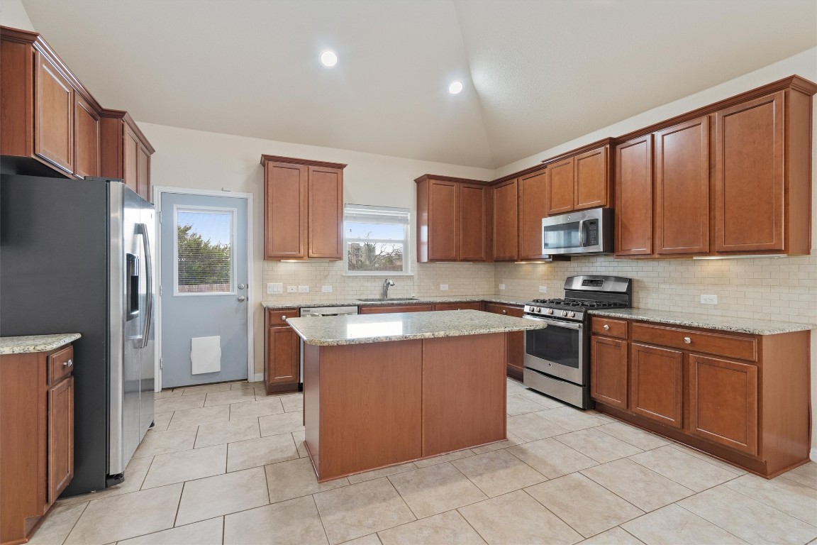 533 Jess Maynard Trail Leander, TX 78641 - Photo 8 of 24 Kitchen with stainless steel appliances, brown cabinetry, light stone counters, lofted ceiling, and a center island