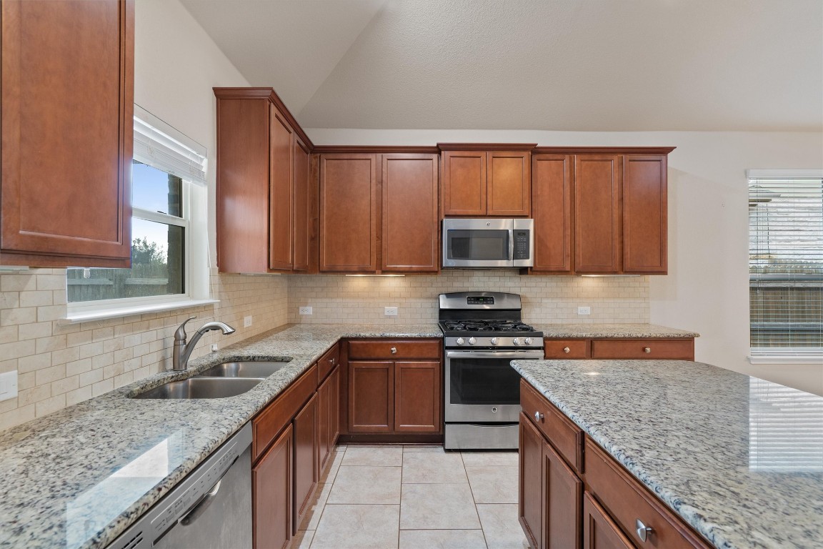 533 Jess Maynard Trail Leander, TX 78641 - Photo 9 of 24 Kitchen with appliances with stainless steel finishes, light stone counters, brown cabinetry, lofted ceiling, and light tile patterned floors