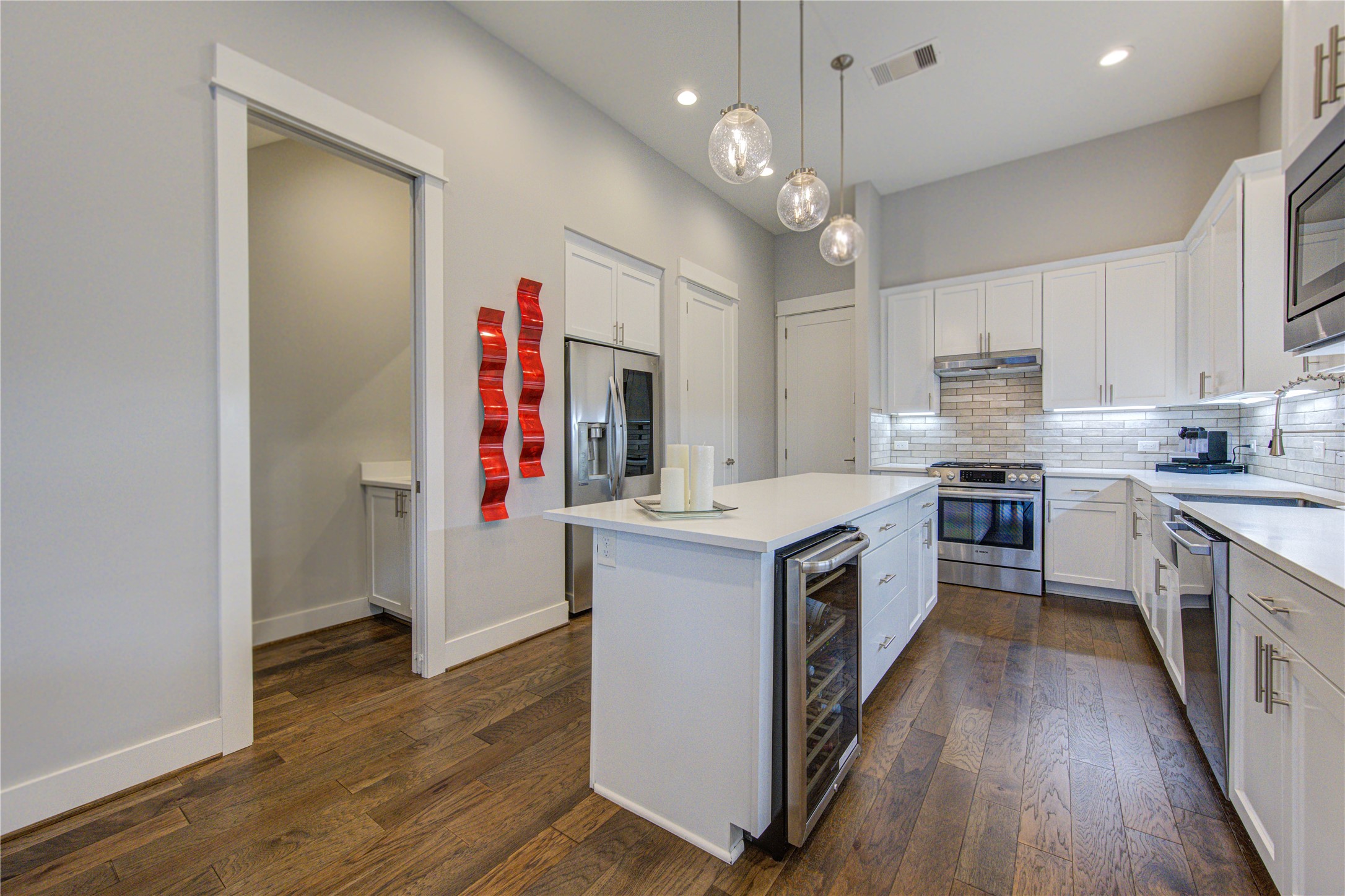 419 Malone Street Houston, TX 77007 - Photo 11 of 47 Modern kitchen featuring white cabinetry, stainless steel appliances, and a central island with pendant lighting. The space has hardwood floors, a stylish backsplash, and a wine fridge, creating a sleek and inviting atmosphere.