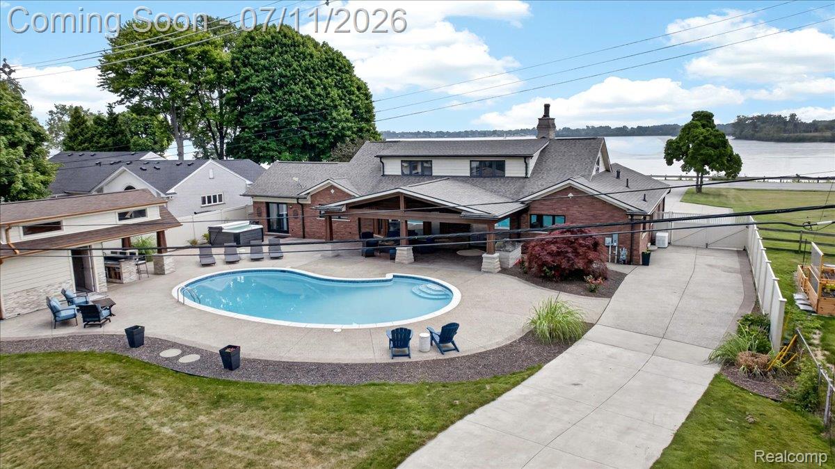 23750 East River Road Grosse Ile, MI 48138 - Photo 43 of 54 Expansive rear yard featuring an irregular-shaped in-ground pool surrounded by concrete decking, a covered outdoor living area with wood beams, a brick and siding exterior, a separate pool house with an outdoor kitchen, and a paved driveway leading to a waterfront area