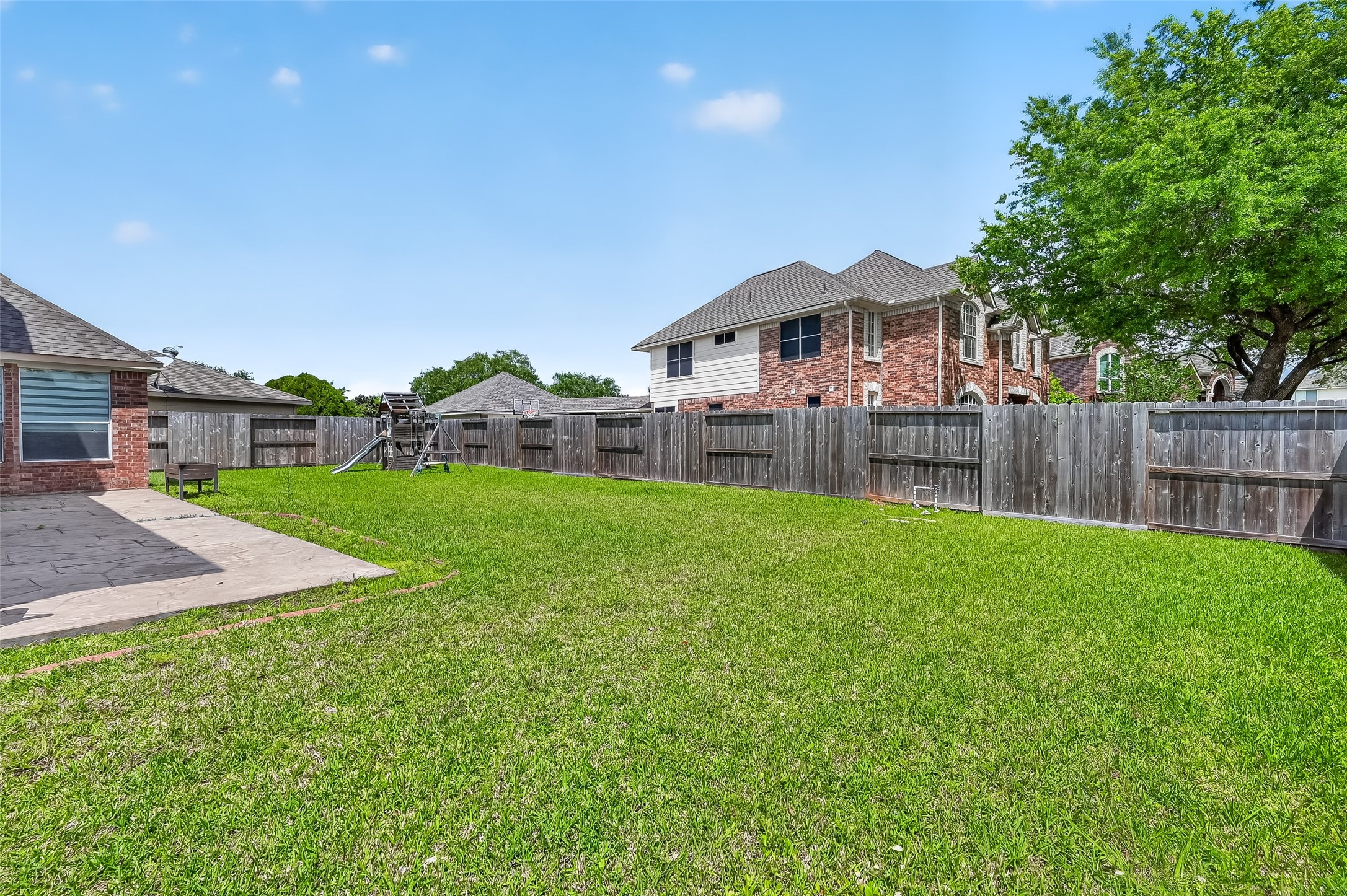 1311 Rene Court Sugar Land, TX 77479 - Photo 43 of 45 This photo showcases a spacious backyard with a well-maintained lawn, bordered by a wooden fence. There's a concrete patio area, suitable for outdoor seating or entertaining, and a children's playset is visible, making it family-friendly. The surrounding houses give a sense of a residential neighborhood.