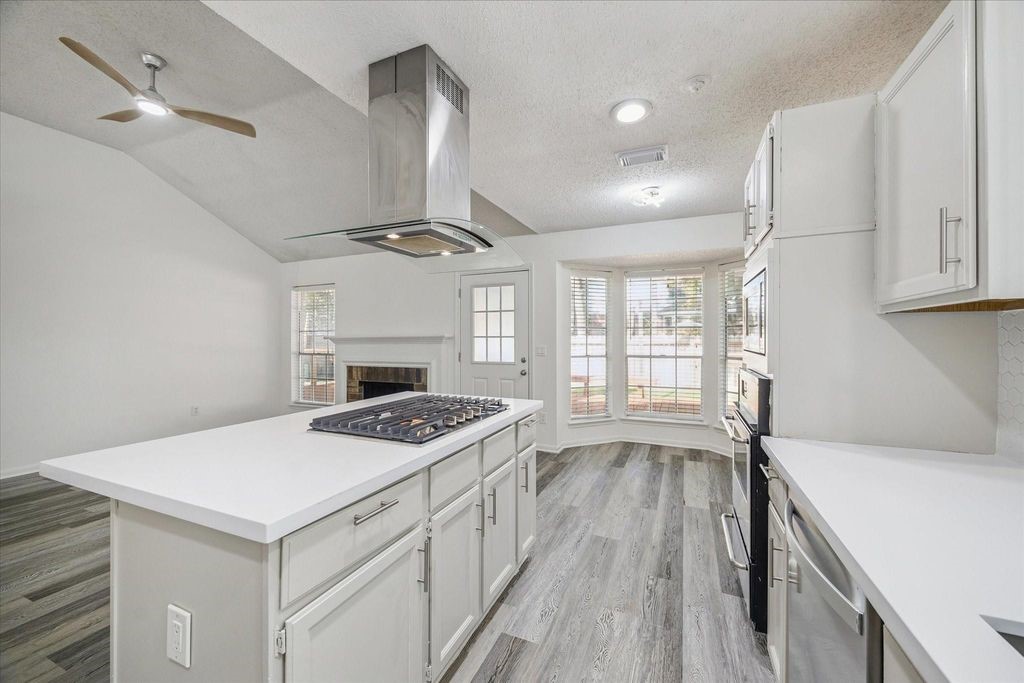143 West Village Knoll Circle Spring, TX 77381 - Photo 14 of 27 This angle highlights the spacious island with a gas cooktop and modern vent hood, overlooking the open living area with high ceilings and fireplace. Natural light floods through the bay windows in the breakfast nook, creating a bright and inviting space that flows seamlessly to the outdoor area.