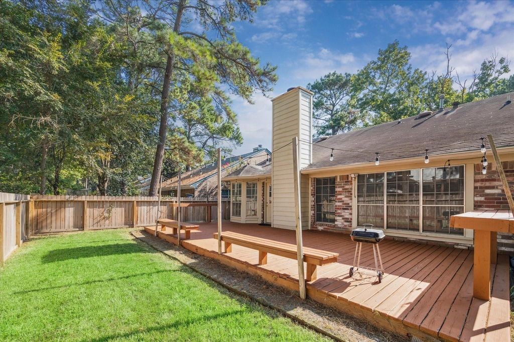 143 West Village Knoll Circle Spring, TX 77381 - Photo 24 of 27 Enjoy outdoor living at its best in this spacious backyard retreat, featuring an expansive wooden deck with built-in seating and charming string lights—perfect for entertaining or relaxing under the trees. Surrounded by mature landscaping and a fully fenced yard, this private outdoor space offers both shade and serenity.