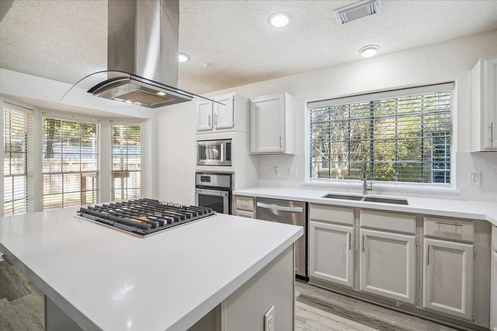 143 West Village Knoll Circle Spring, TX 77381 - Photo 9 of 27 This modern kitchen is a dream, featuring sleek quartz countertops, a stylish glass range hood, a gas cooktop island, and stainless steel appliances—including a built-in microwave, oven, and French-door refrigerator. Crisp white cabinetry and a classic tile backsplash complete the bright, clean look.