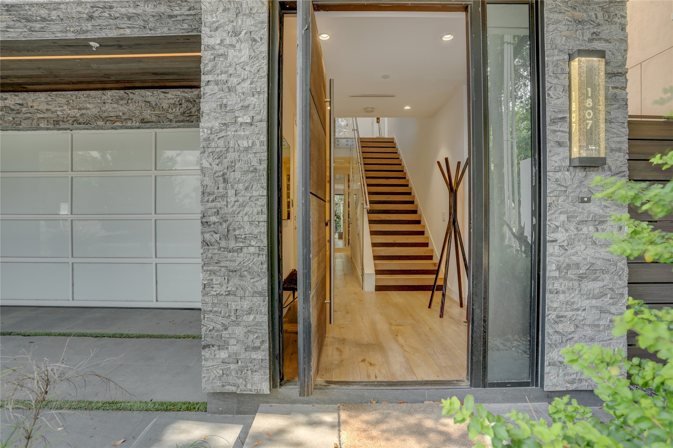 1807 Indiana Street Houston, TX 77006 - Photo 1 of 35 Grand first-floor entry featuring an oversized pivoting wood door, modern stacked stone accent walls, sleek frosted glass garage doors, and wide-plank flooring leading to the staircase.