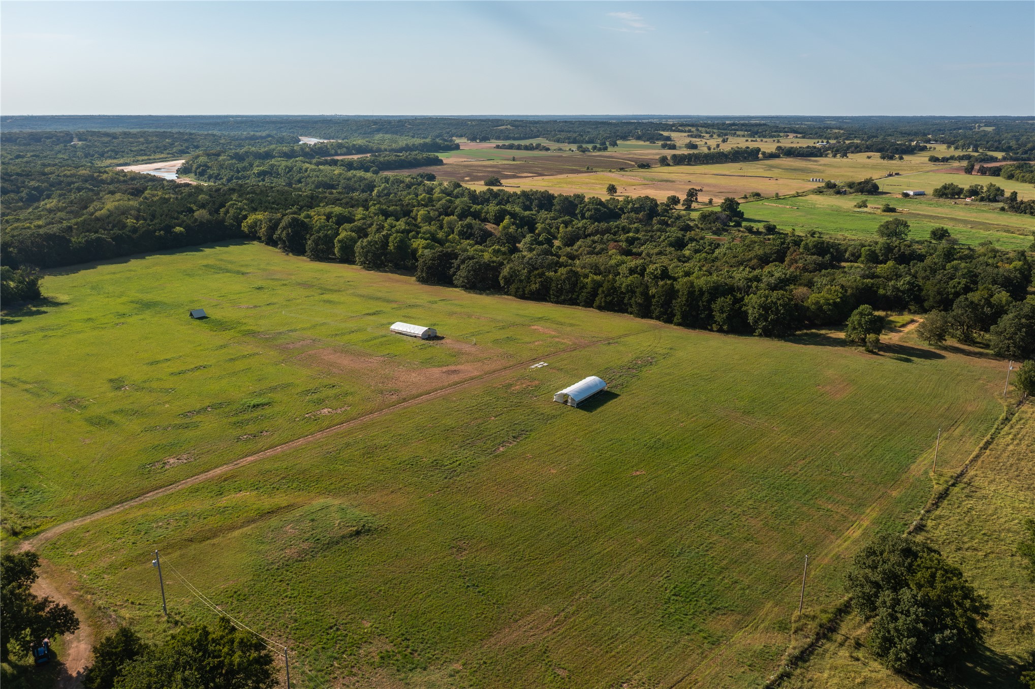 0 West Dunkin Road Cushing, OK 74023 - Photo 3 of 62 Expansive green fields, two white outbuildings, dense tree lines, distant river, and a clear sky