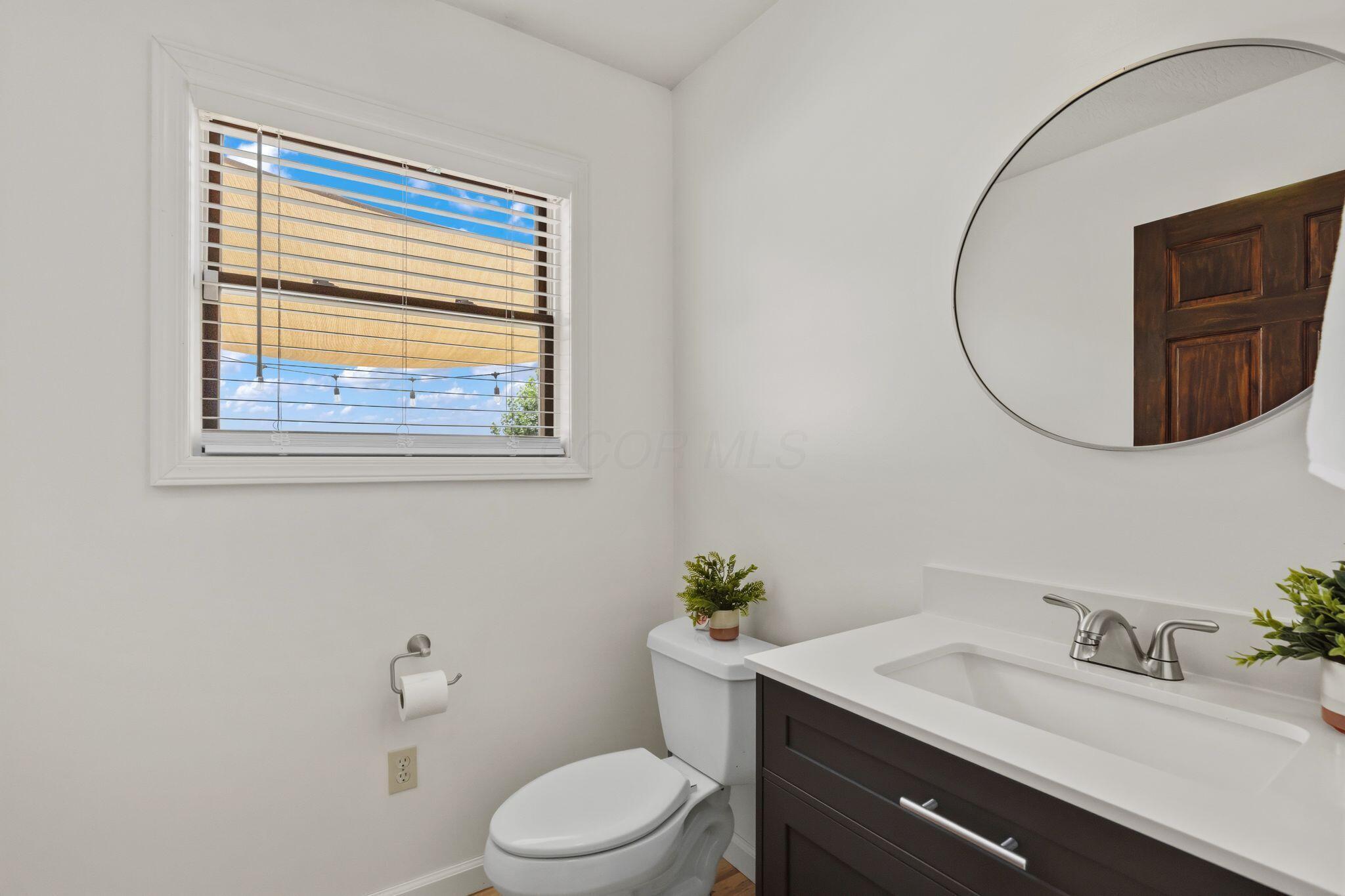 10446 Rich Hollow Road Southwest Lancaster, OH 43130 - Photo 21 of 83 Bathroom featuring a white vanity with a dark wood-finish cabinet and integrated sink, a round wall-mounted mirror, a window with horizontal blinds, and a white toilet