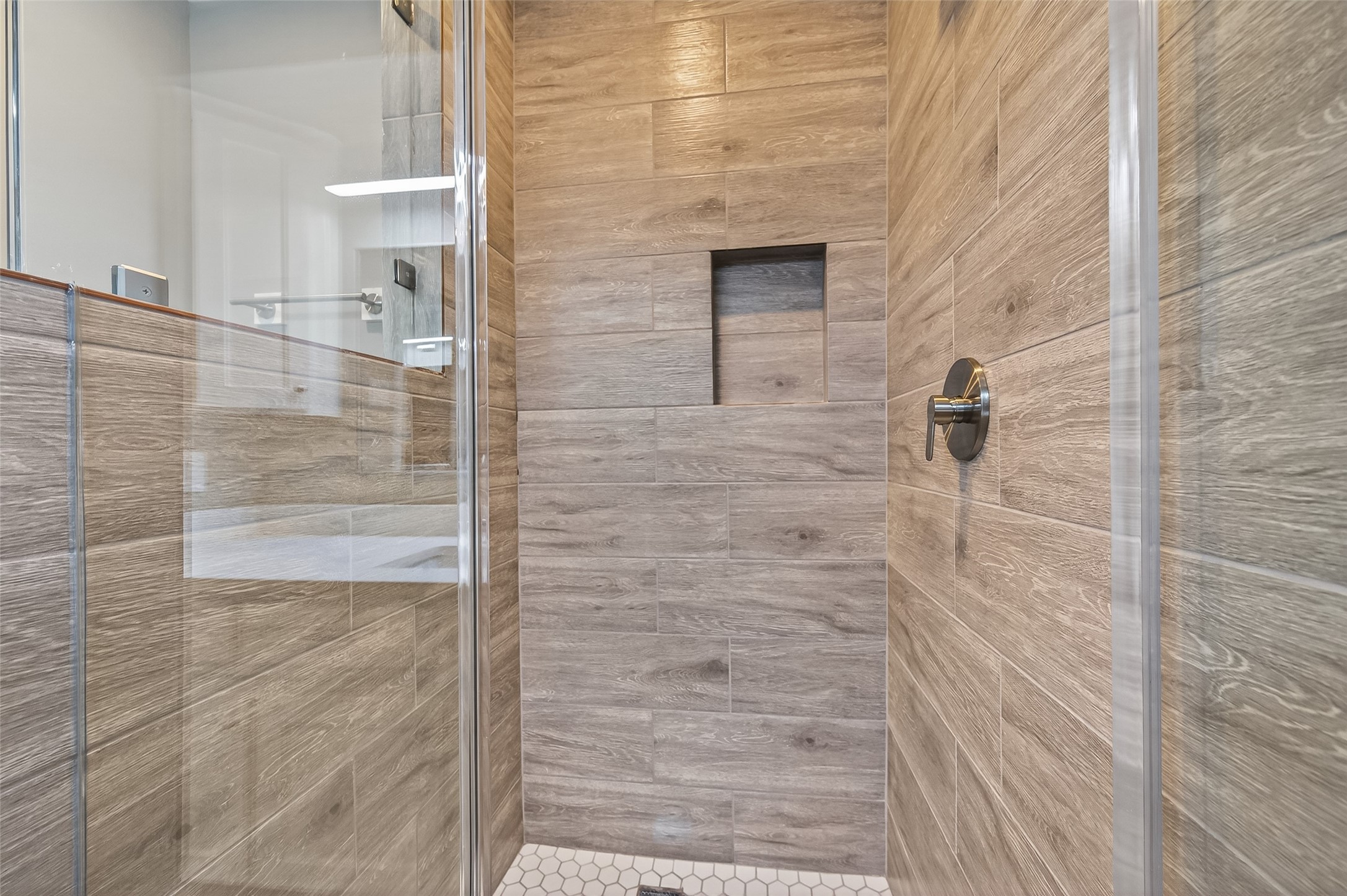 902 Kingsbridge Road Houston, TX 77073 - Photo 36 of 43 This contemporary primary shower features large-format, wood-look wall tile with a convenient recessed storage niche. The space is finished with a frameless glass enclosure, a sleek brushed-metal shower handle, and classic white hexagonal mosaic floor tile.
