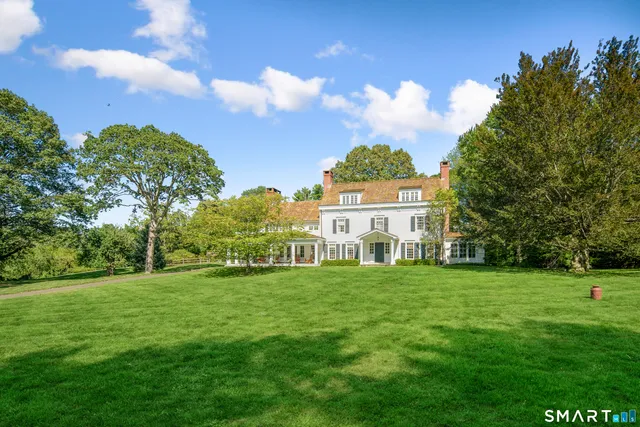 a view of a white house in front of a big yard with plants and large trees