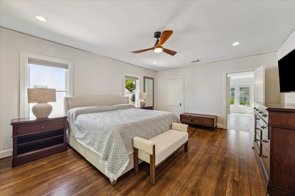 1925 Banks Street Houston, TX 77098 - Photo 17 of 38 A spacious and bright primary bedroom with hardwood floors, neutral walls, and recessed lighting. Features a ceiling fan, large bed, and ample furniture, including nightstands and a dresser. Two windows provide natural light, and there is an adjoining room visible.