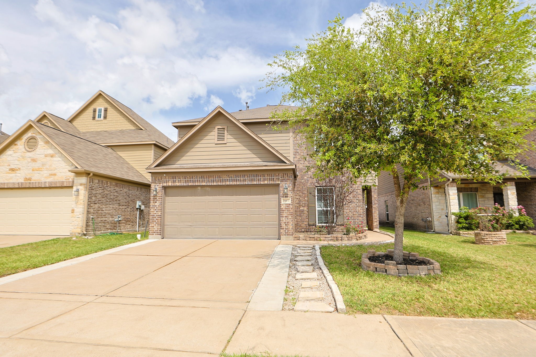 This photo showcases a two-story brick home with a double garage, a well-maintained front yard, and a mature tree. The driveway is spacious, and the house has a charming, welcoming exterior.