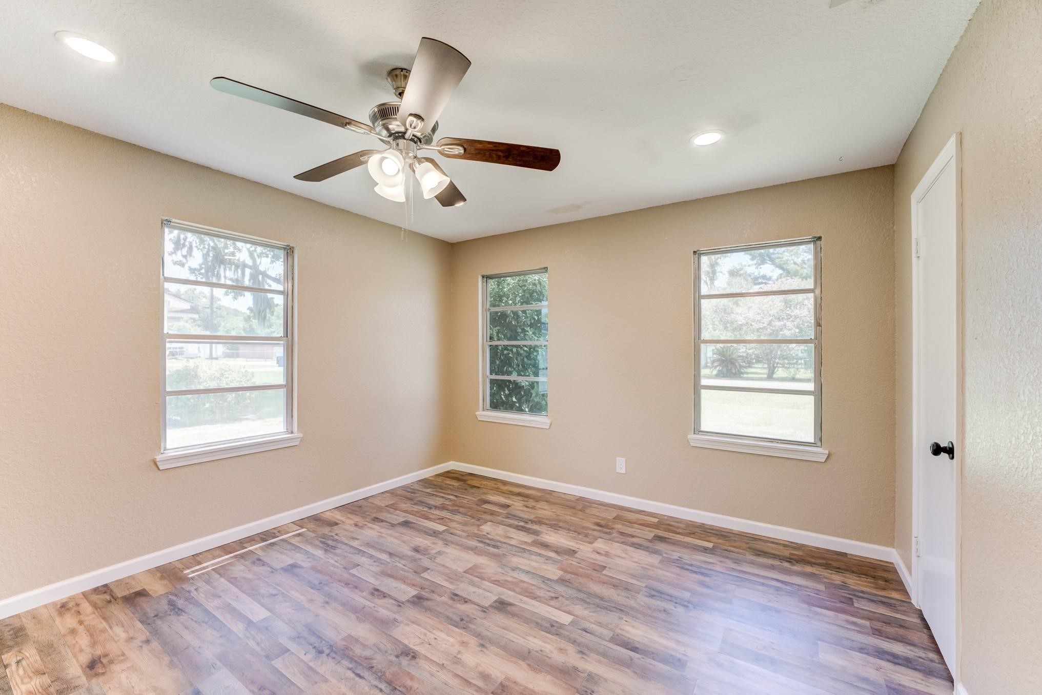 305 Yaupon Street Sweeny, TX 77480 - Photo 5 of 22 Bright room with three windows, neutral walls, and wood-look flooring. Features a ceiling fan with lights, creating a cozy and airy atmosphere.