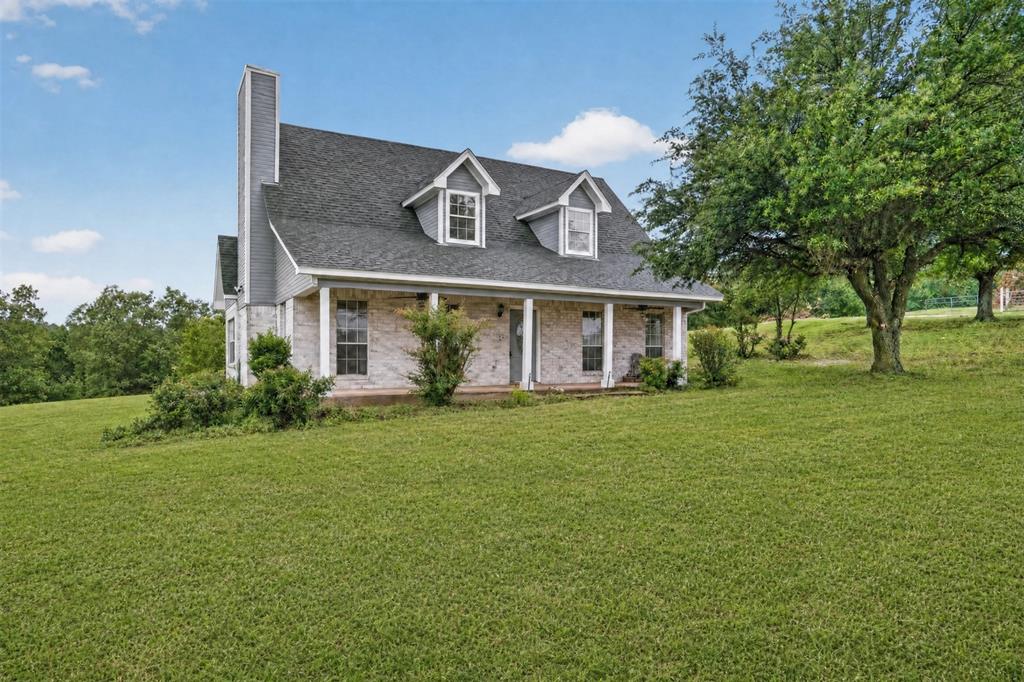 Virtually enhanced. White brick exterior with a dark shingle roof, twin dormer windows, and a covered front porch featuring white columns