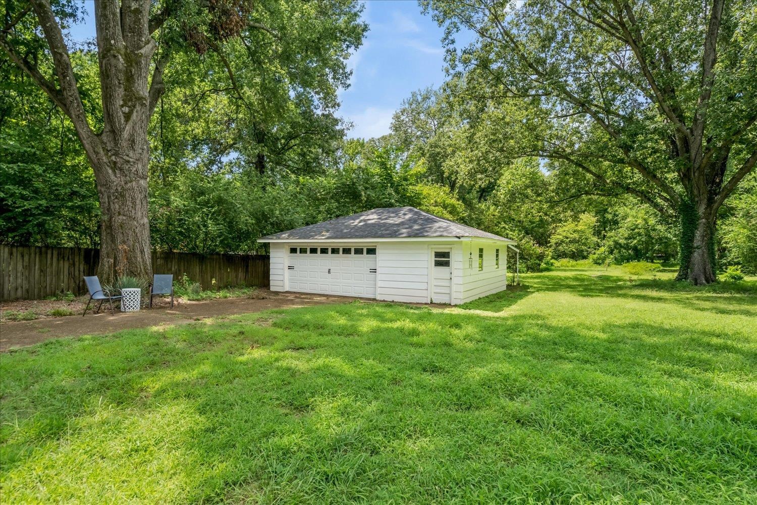 Detached two-car garage with white siding, surrounded by mature trees and a wooden privacy fence