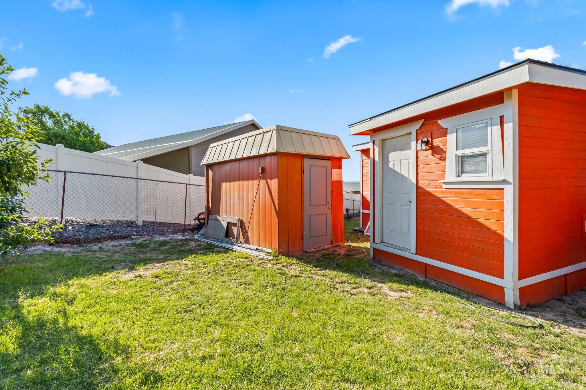 20106 Lower Pleasant Ridge Road, Unit 29 Caldwell, ID 83607 - Photo 18 of 19 Expansive green lawn featuring a vibrant orange outbuilding with white trim, a secondary brown shed with a light brown roof, and a combination chain-link and vinyl privacy fence