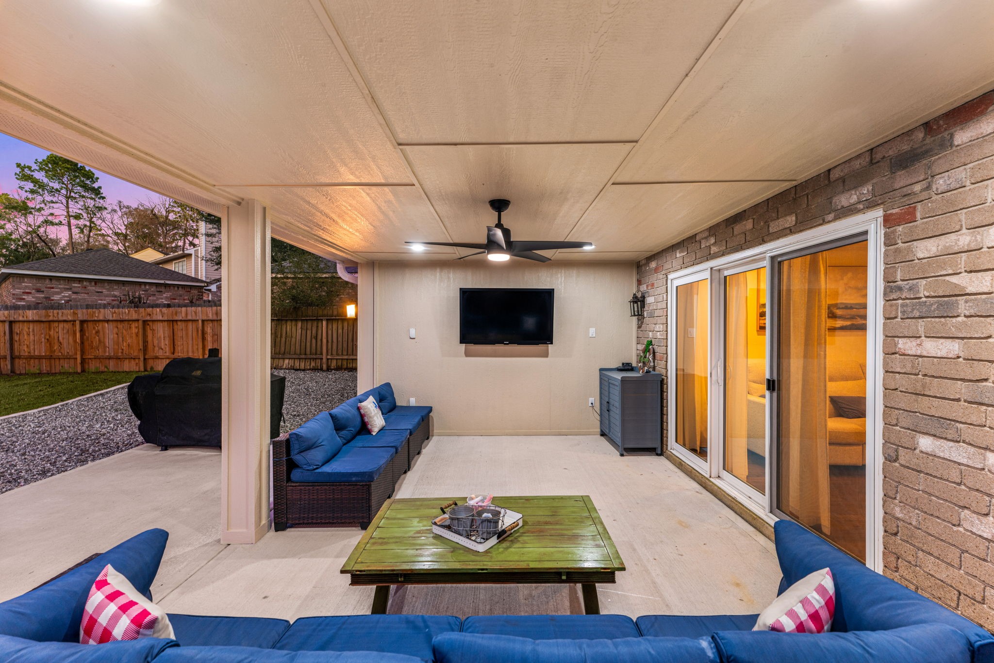 57 Fire Flicker Place Spring, TX 77381 - Photo 5 of 11 Outdoor living at its funnest under this covered patio with comfortable seating, ceiling fan & wall-mounted TV(TV not included). This space is perfect for outdoor entertainment, featuring a brick wall and sliding glass doors that lead inside. The area also includes a neatly landscaped yard with privacy fencing.