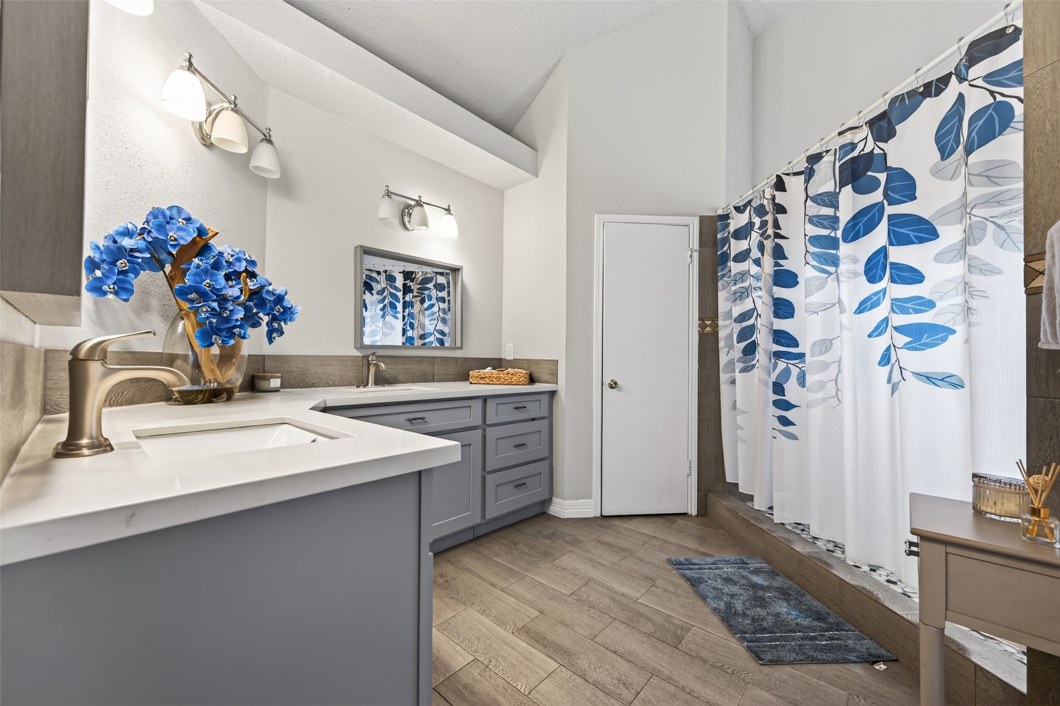 18446 North Willow Bluff Road Katy, TX 77449 - Photo 21 of 33 This modern bathroom features a spacious layout with vaulted ceilings, a double vanity with sleek gray cabinets, and elegant fixtures. The blue and white decor, including a leaf-patterned shower curtain and vibrant floral arrangement, adds a refreshing touch.