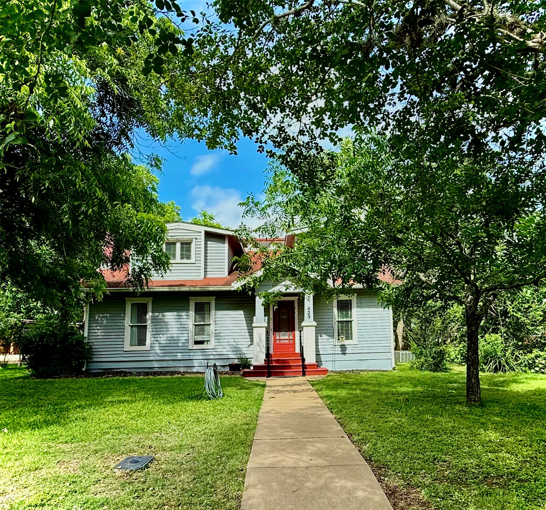 423 South Main Street Lockhart, TX 78644 - Photo 1 of 26 Traditional light blue siding residence featuring a red front door, white trim, red shingled roof, a paved walkway, and mature trees