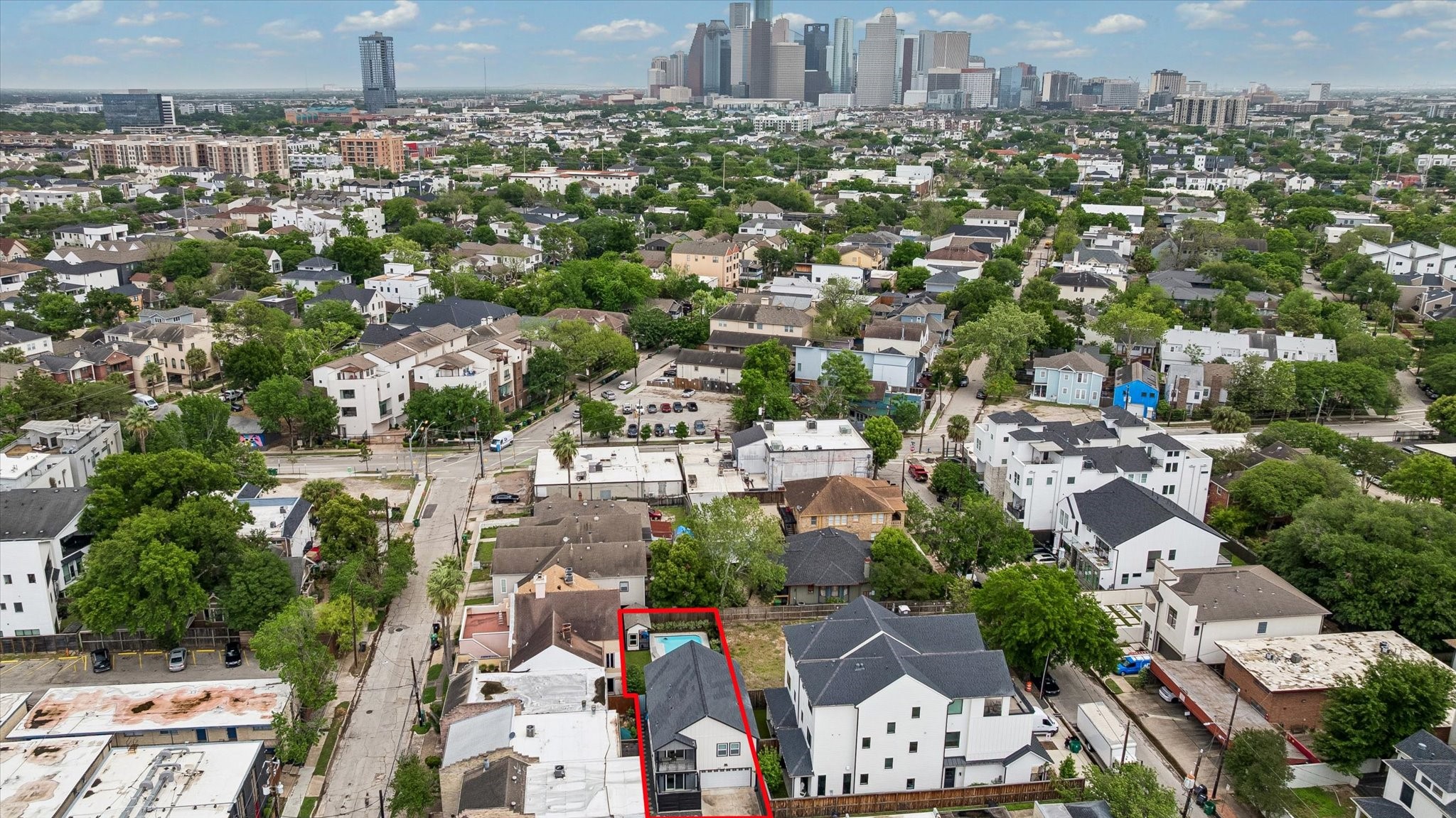 1420 Indiana Street Houston, TX 77006 - Photo 30 of 31 This aerial photo showcases a residential neighborhood with a modern home outlined in red. The area is densely populated with a mix of contemporary and traditional houses, abundant greenery, and tree-lined streets. The city skyline is visible in the background, indicating proximity to urban amenities.