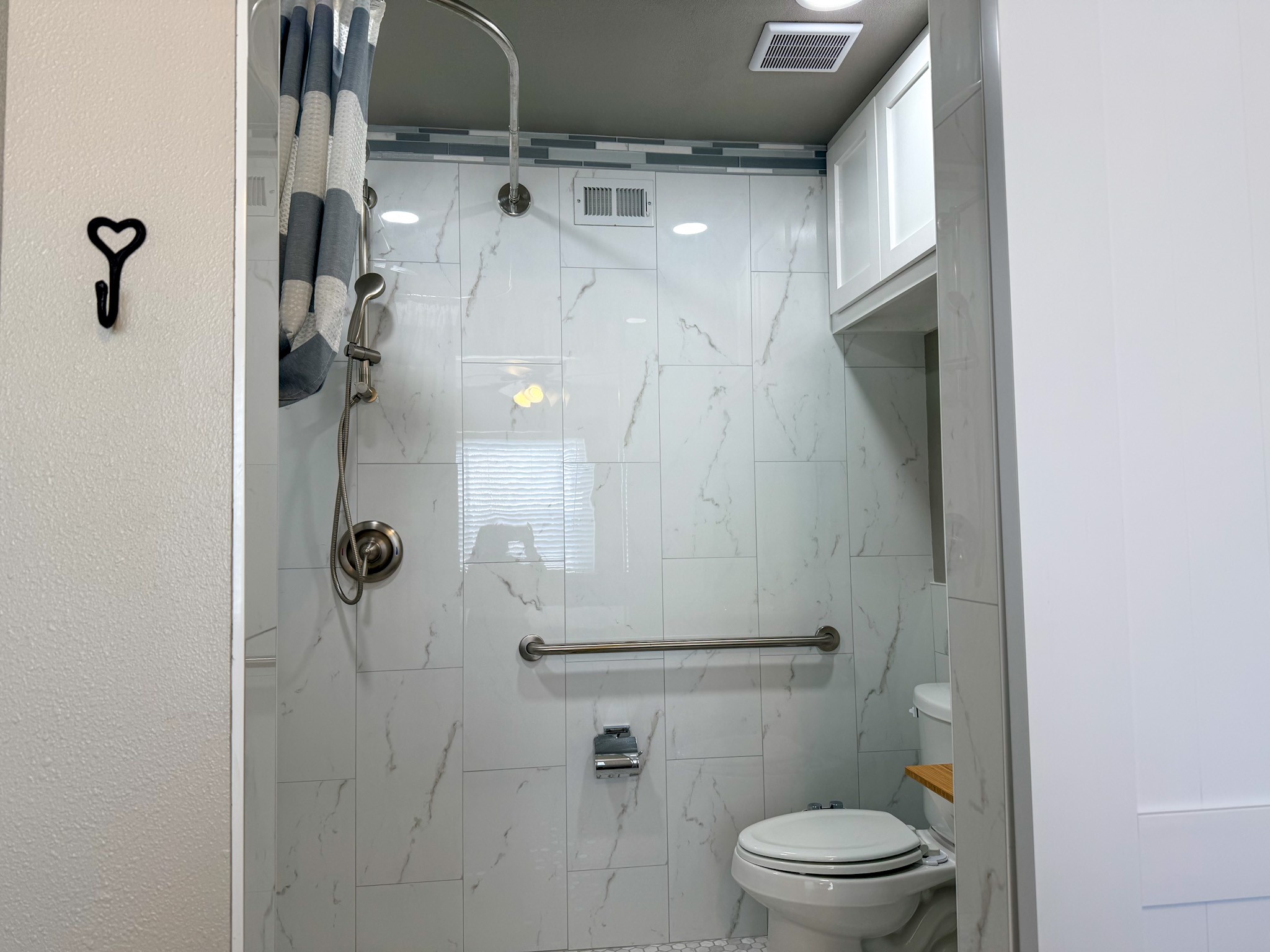24527 Appian Ridge, Unit B Porter, TX 77365 - Photo 9 of 10 This bathroom features a modern walk-in shower with marble-style tiles, a handheld showerhead, and a grab bar for safety. The space includes a white toilet, upper storage cabinets, and a striped shower curtain for a clean, functional design.