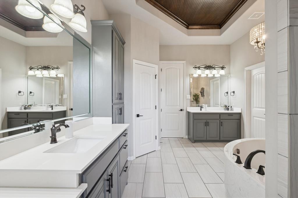 542 Pierce Road Red Oak, TX 75154 - Photo 17 of 27 Spacious bathroom featuring dual vanities with white countertops and dark cabinetry, a built-in soaking tub with dark fixtures, light tile flooring, and a tray ceiling with a wood-paneled accent