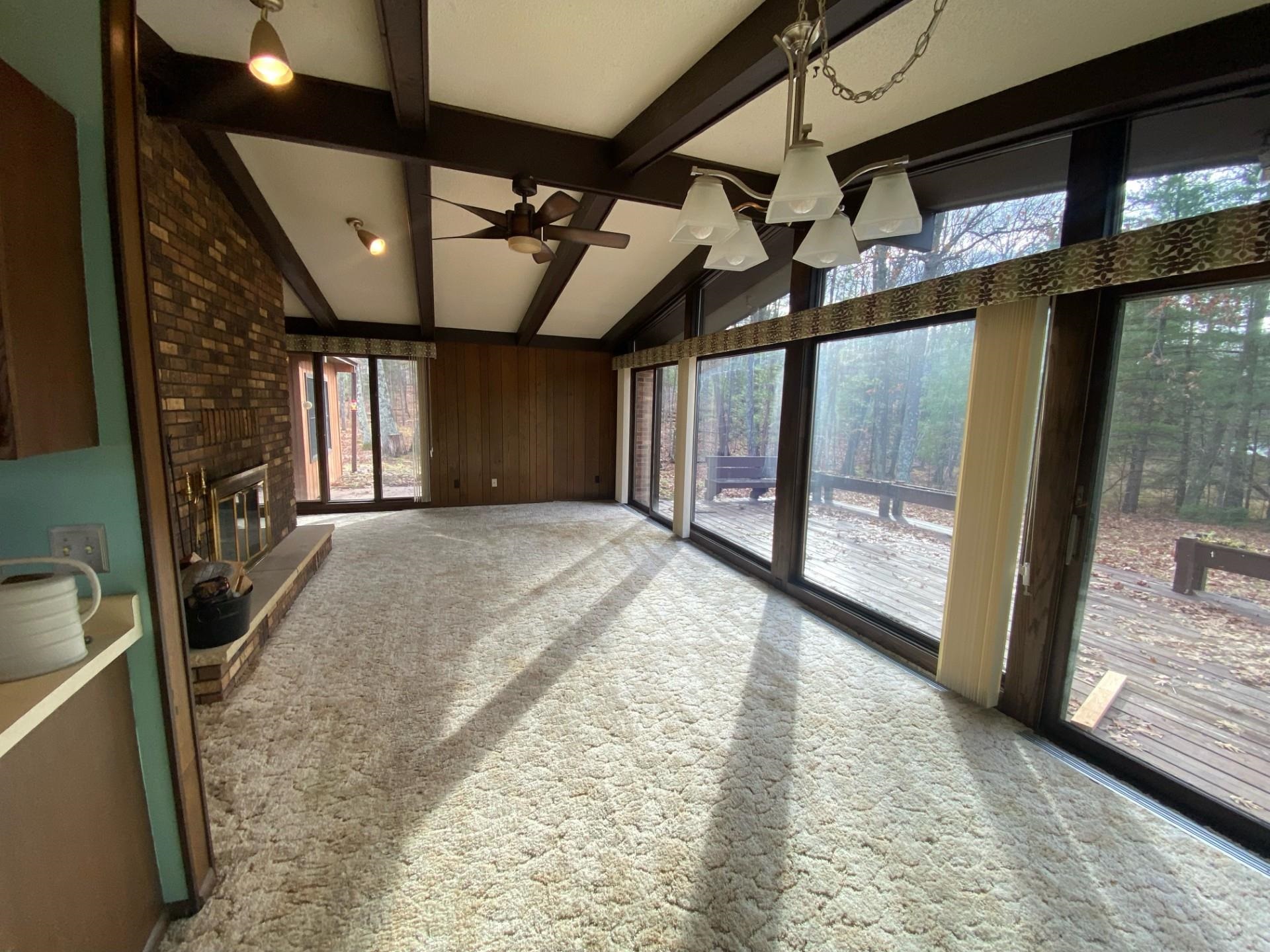 11316 East Timber Ridge Drive Lake Nebagamon, WI 54849 - Photo 7 of 27 Unfurnished living room with ceiling fan with notable chandelier, carpet, vaulted ceiling with beams, a brick fireplace, and wooden walls