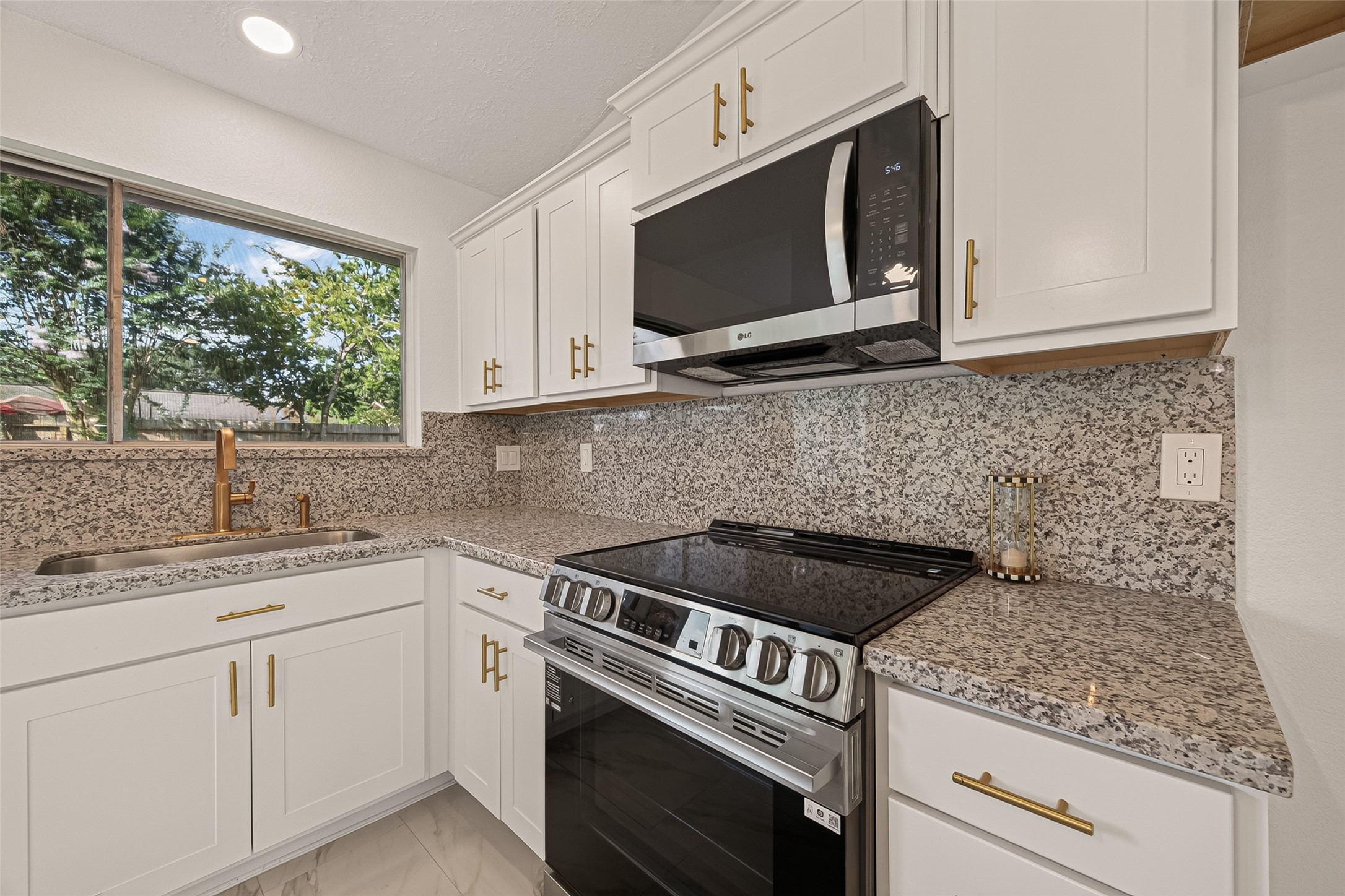16619 Tibet Road Friendswood, TX 77546 - Photo 7 of 29 This modern kitchen features sleek white cabinetry, granite countertops, and stainless steel appliances, including a built-in microwave and stove. A large window provides natural light, and there are stylish gold fixtures and handles.