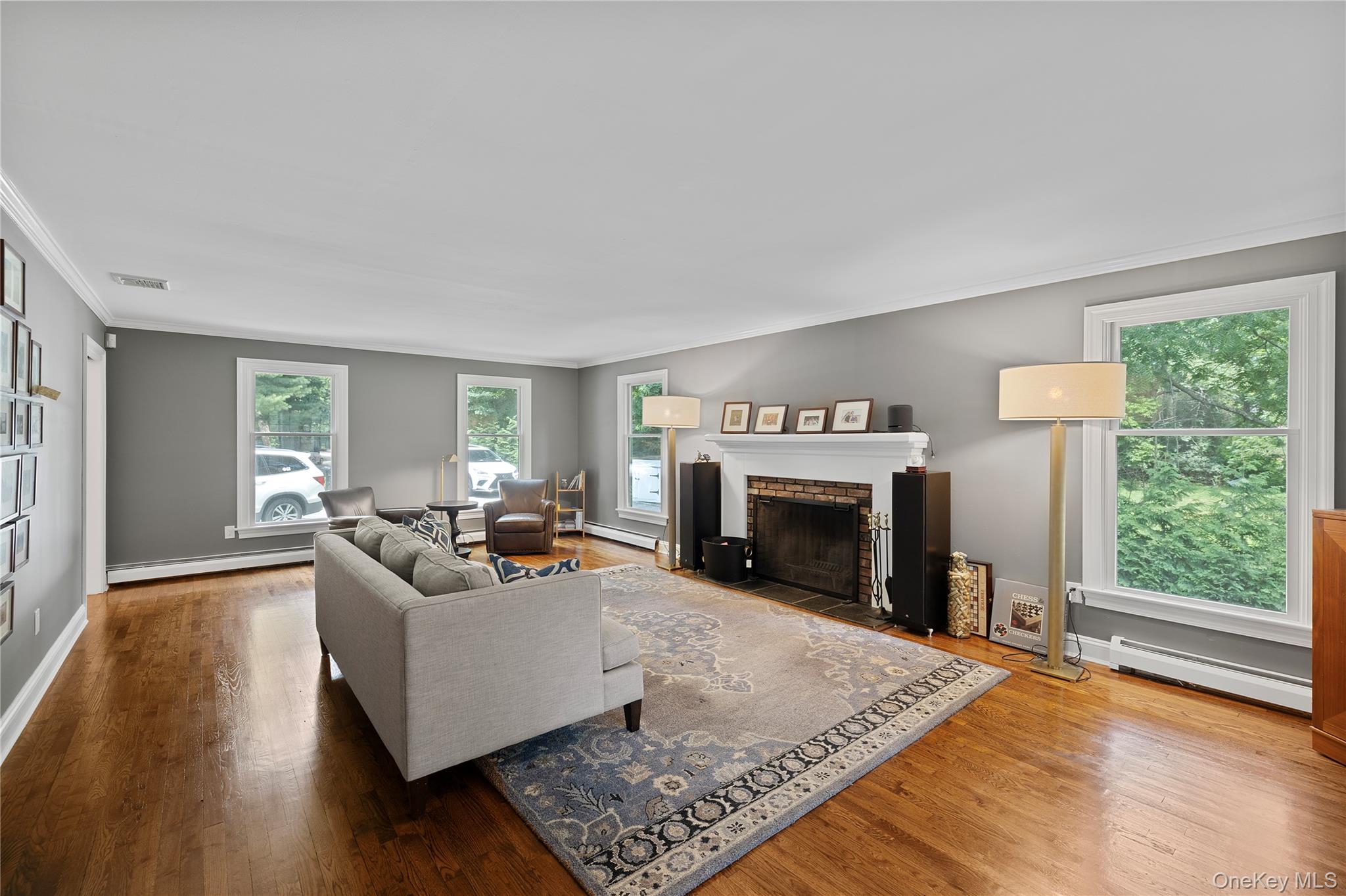 9 Three Sisters Road St. James, NY 11780 - Photo 2 of 36 Spacious living room featuring hardwood flooring, a white mantel wood burning fireplace with brick surround, multiple windows, and crown molding