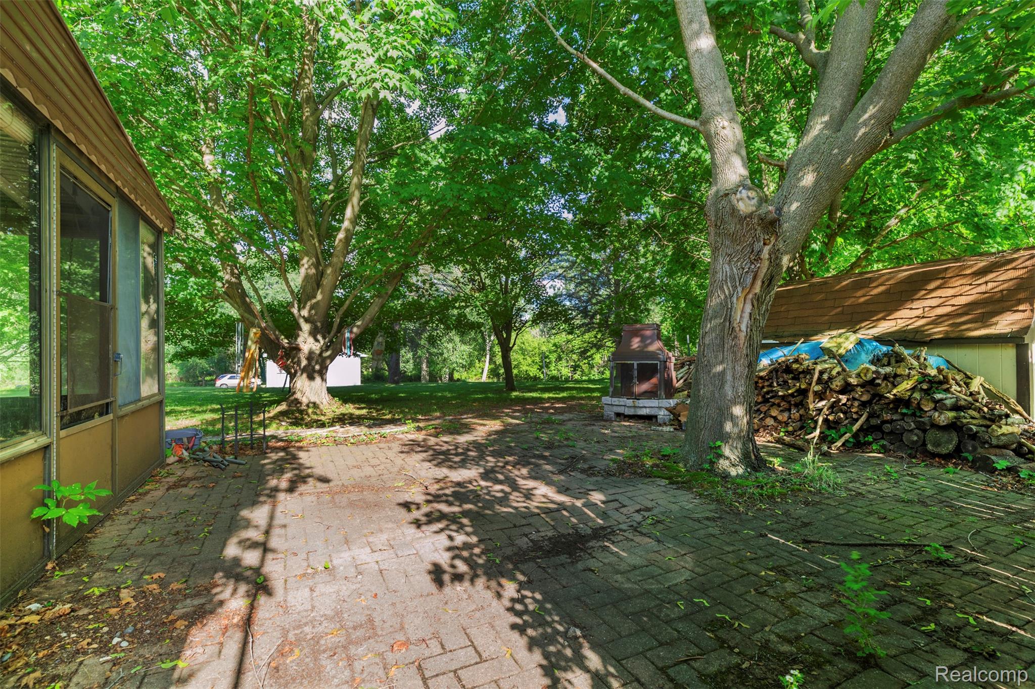 309 Windy Bluff Street Flushing, MI 48433 - Photo 27 of 32 Expansive brick paver patio area surrounded by mature trees, featuring a freestanding outdoor fireplace and a screened-in porch with multiple windows