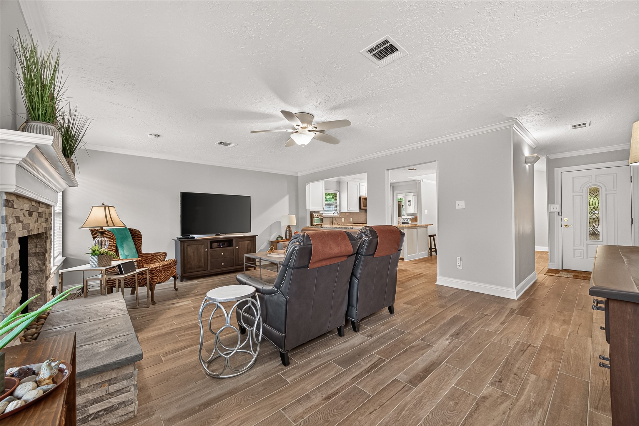 24719 Wilderness Road Spring, TX 77380 - Photo 13 of 42 This inviting living room features a cozy stone fireplace, elegant ceramic tile flooring, and an open layout leading to a modern kitchen. The space is well-lit and includes a ceiling fan, making it perfect for relaxing or entertaining.