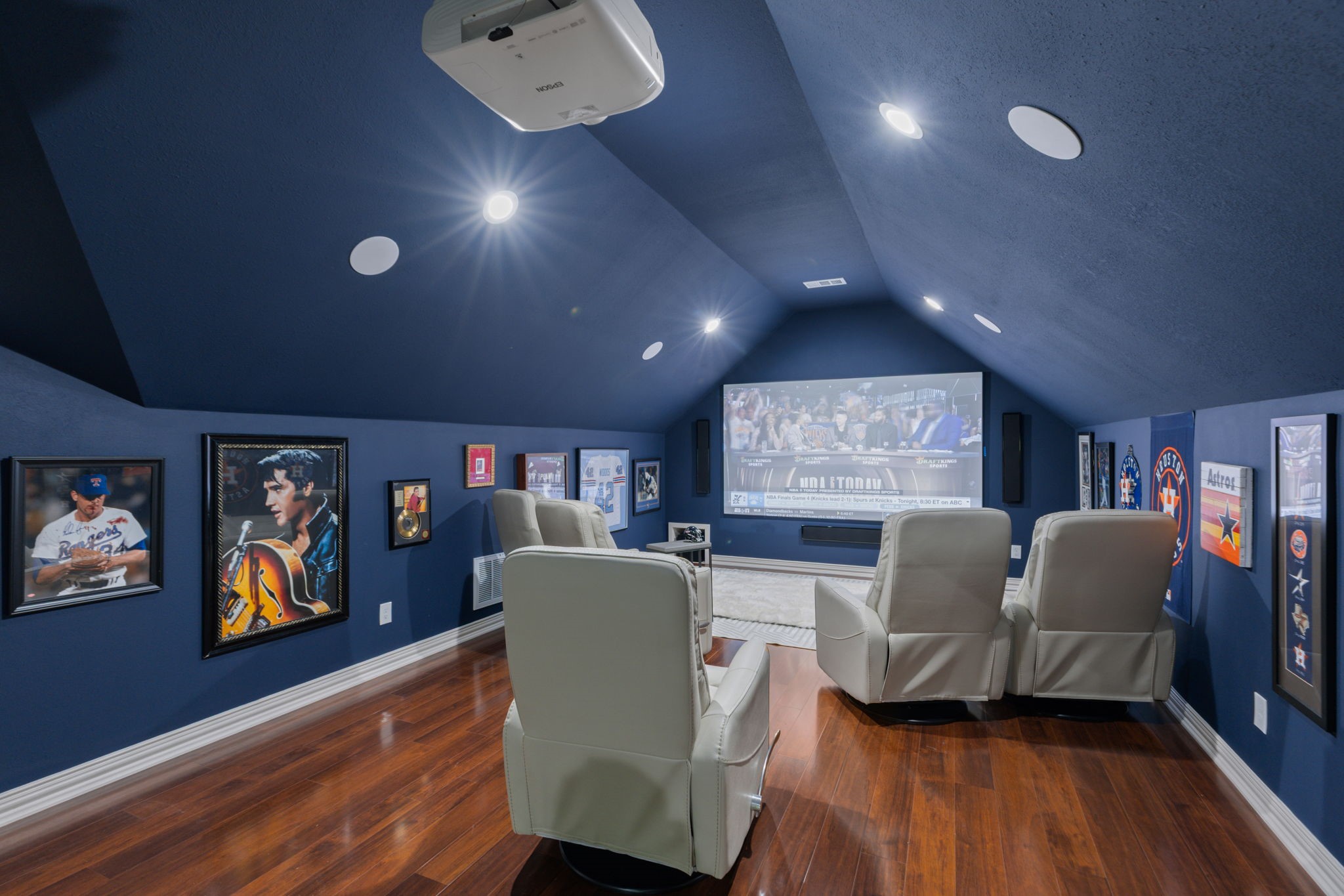 39 Maymont Way Spring, TX 77382 - Photo 35 of 50 This photo showcases a cozy home theater room with dark blue walls and hardwood floors. It features a large screen, ceiling-mounted projector, and four plush recliners. The room is decorated with sports memorabilia, providing a stylish and private entertainment space.