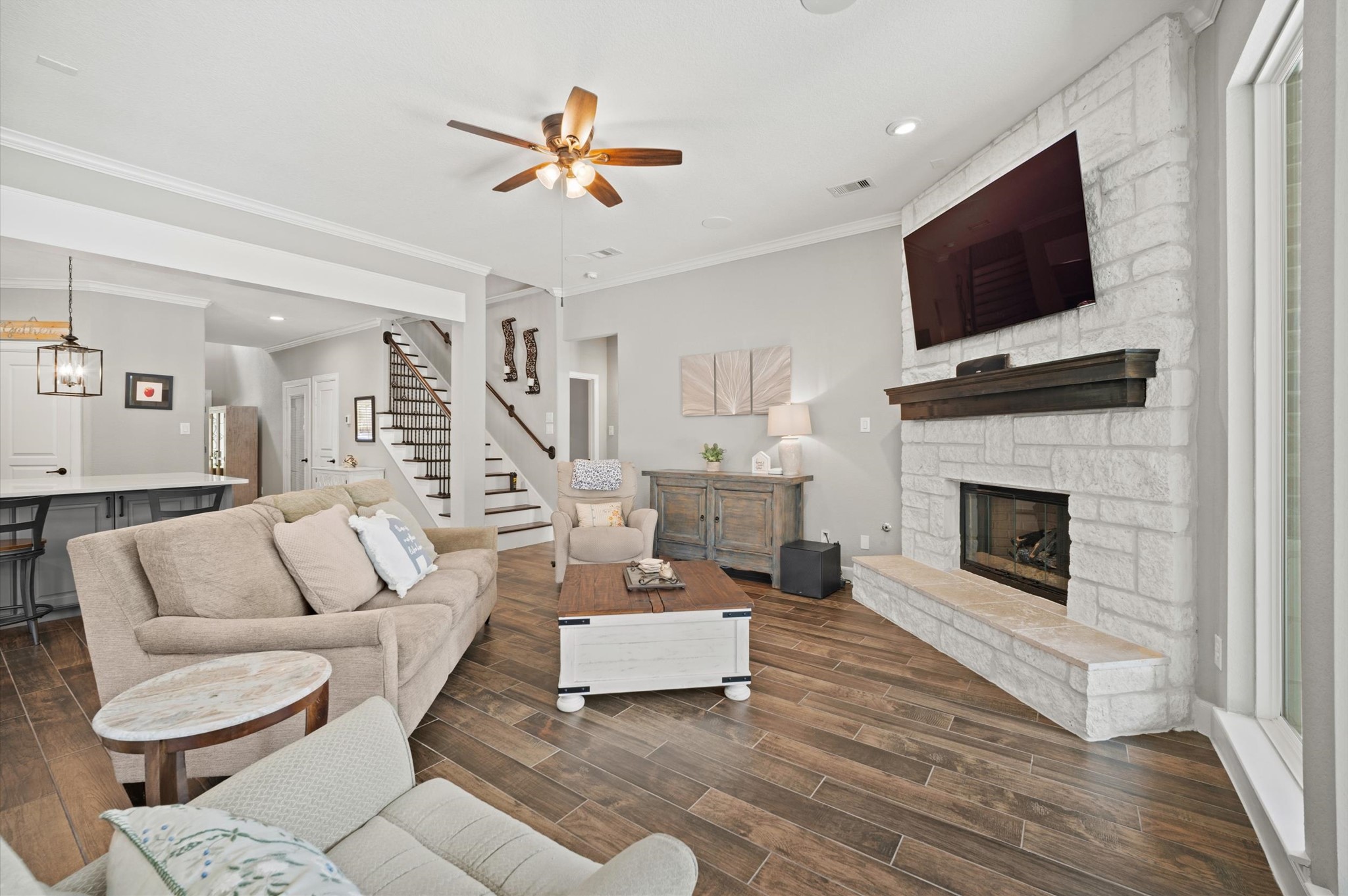 9507 Empress Crossing Drive Spring, TX 77379 - Photo 18 of 50 This inviting living room features an open layout with wood-like tile flooring, a cozy stone fireplace with a mounted TV, and ample seating. It seamlessly connects to a modern kitchen, visible in the background, and offers a warm, elegant atmosphere perfect for relaxation and entertainment.