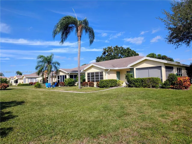 a view of a white house in front of a big yard with plants and large trees
