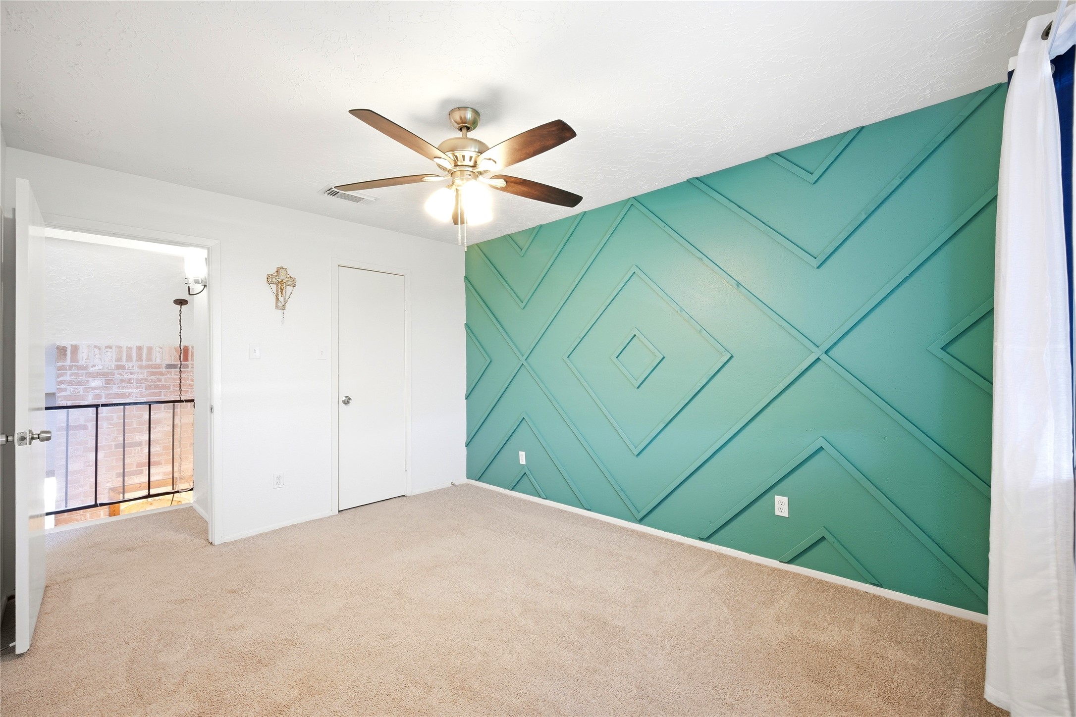 4718 Temple Bell Drive Spring, TX 77388 - Photo 30 of 50 This bright room features a unique geometric accent wall in teal, complemented by neutral carpeting. It includes a ceiling fan, a window with curtains, and an entry door leading to a balcony with a view of an exposed brick wall.