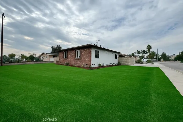 a view of a white house in front of a big yard with plants and large tree