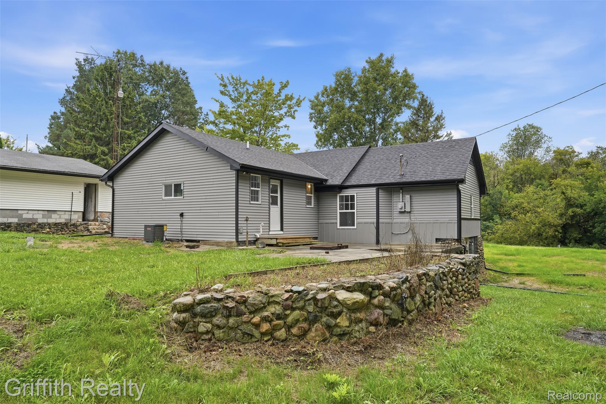 5820 Mack Road Howell, MI 48855 - Photo 31 of 60 Grey siding exterior with a dark shingle roof, accompanied by a stone retaining wall and a concrete patio space