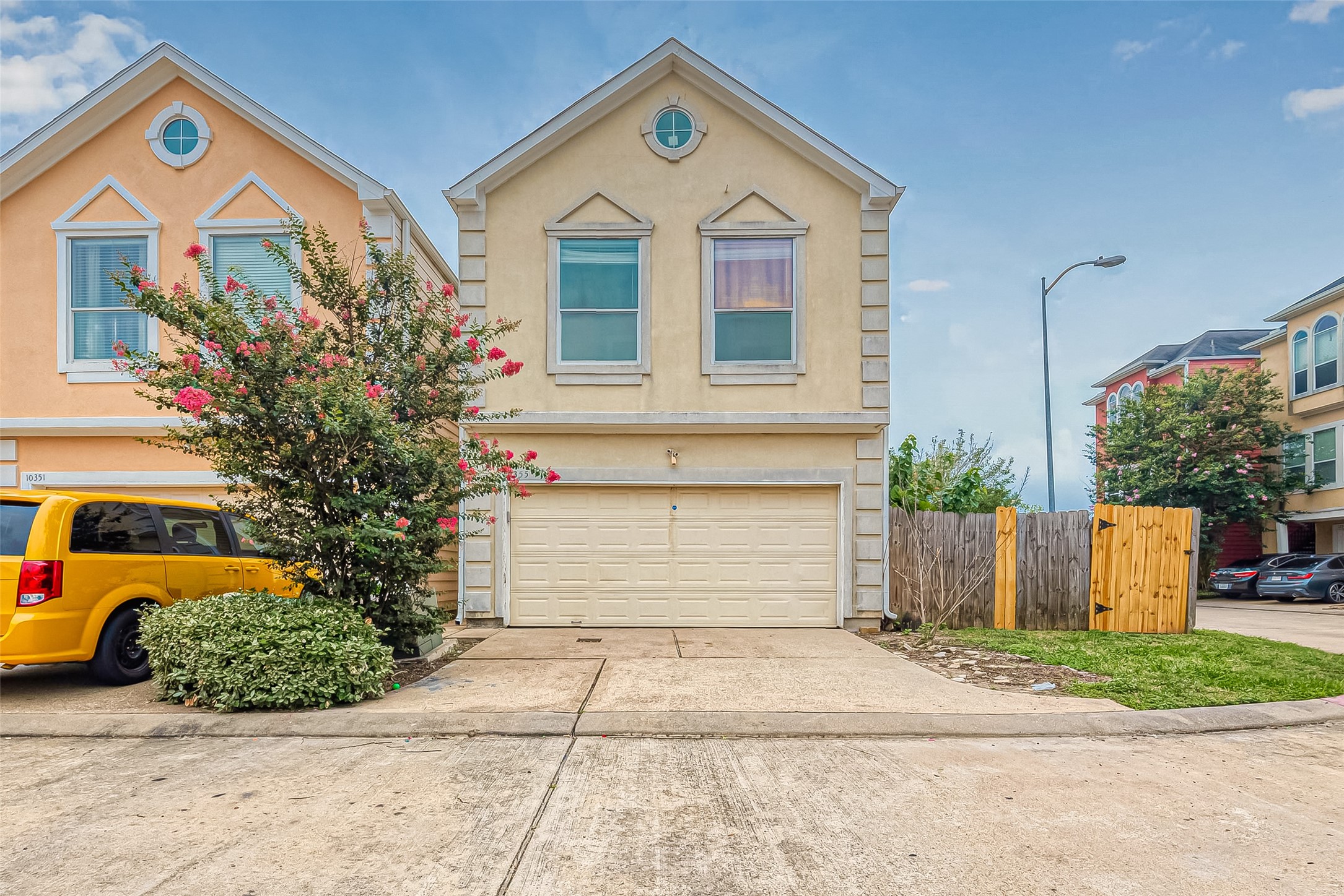 This is a two-story, single-family home with a beige exterior and a two-car garage. It features a small front yard with a flowering tree, and a wooden fence on one side. The neighborhood appears residential, with similar homes nearby.