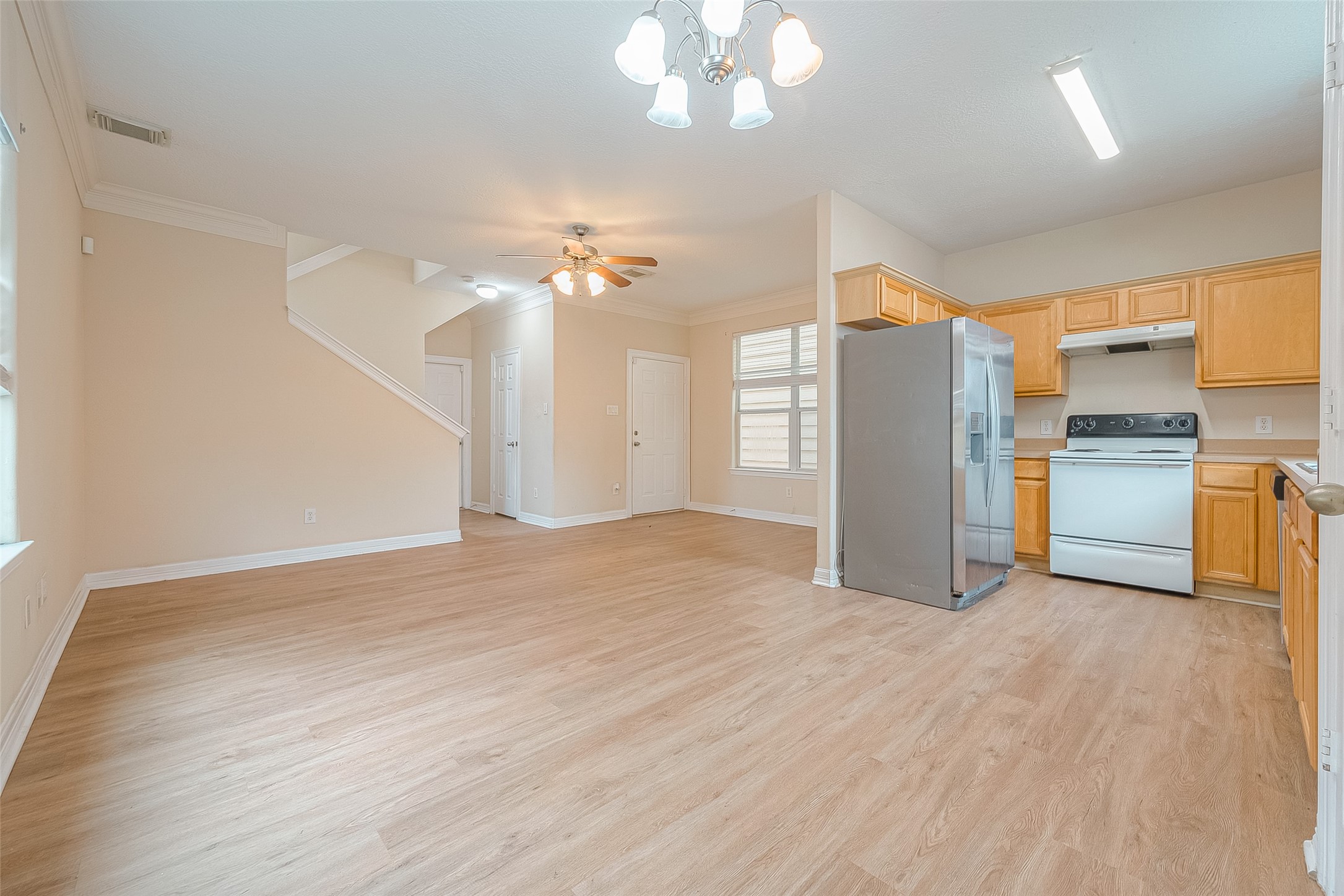 10355 Panther Point Drive Houston, TX 77099 - Photo 15 of 44 This photo showcases a bright, open-concept area with light wood flooring, featuring a kitchen with wooden cabinets, a stainless steel refrigerator, and a white stove. The room includes a dining area with overhead lighting and a ceiling fan, and there's a staircase leading to another level.