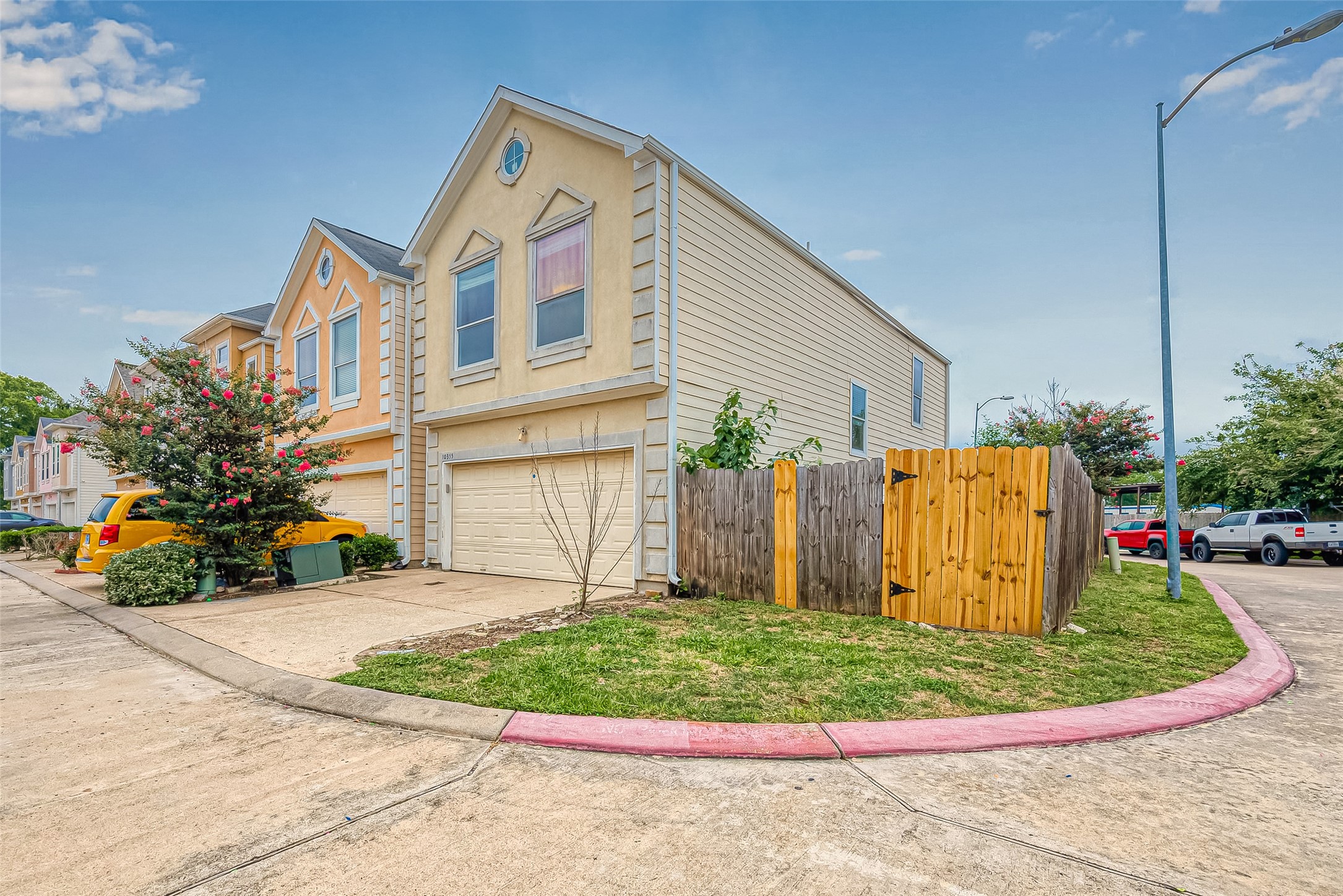 10355 Panther Point Drive Houston, TX 77099 - Photo 3 of 44 This photo shows a two-story, tan townhouse with a gable roof and attached garage. It's located on a corner lot with a small fenced yard. The surrounding area is landscaped with flowering shrubs, and the neighborhood features a row of similar homes.