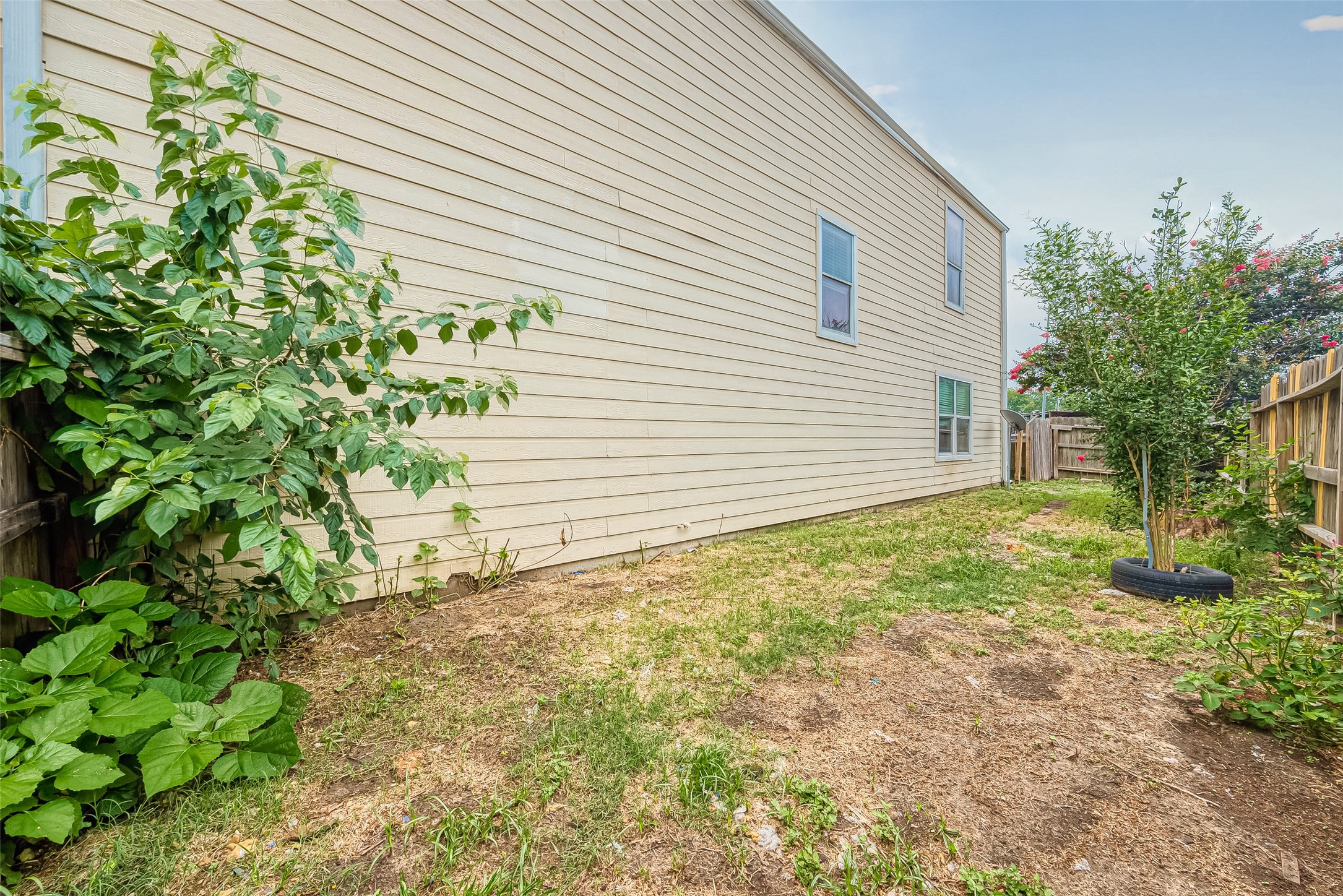 10355 Panther Point Drive Houston, TX 77099 - Photo 39 of 44 This image shows a side yard with a beige siding exterior, window views, and a wooden fence. The yard features a grassy area with some plants and small trees, offering potential for gardening or landscaping.