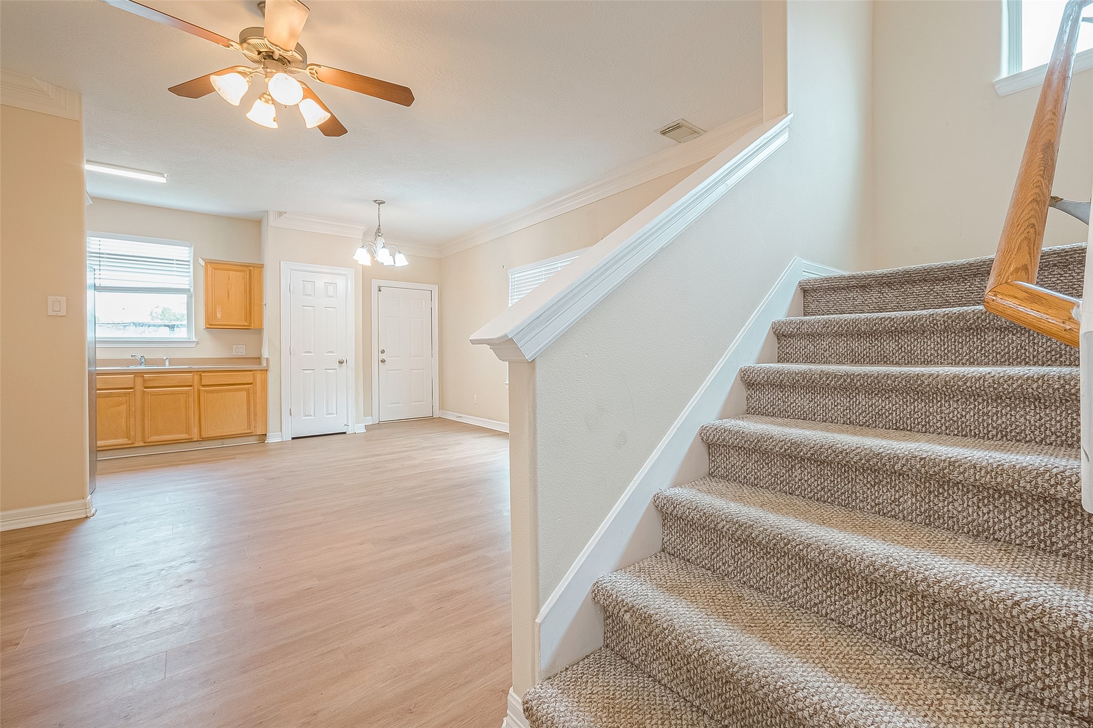 10355 Panther Point Drive Houston, TX 77099 - Photo 9 of 44 This photo features a bright, open-plan area with light wood flooring and a neutral color palette. The space includes a kitchen with wooden cabinets, a dining area with overhead lighting, and carpeted stairs leading to another level. Large windows allow natural light to fill the space.