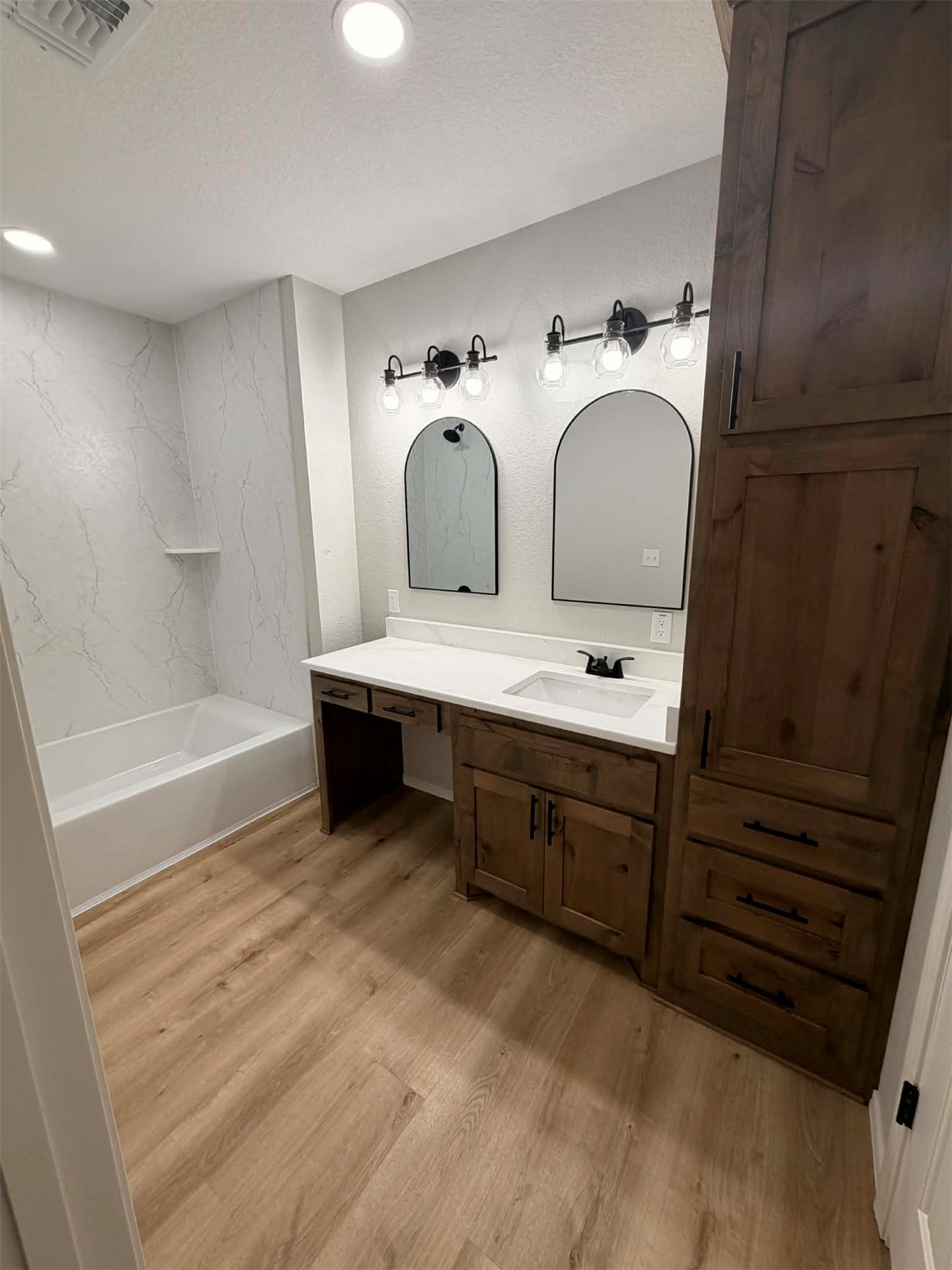 2911 East 2090 Road Hugo, OK 74743 - Photo 13 of 22 Wood-finish flooring, a white built-in tub with a tiled surround, a vanity with a white countertop, dark wood-finish cabinetry, and two arched mirrors