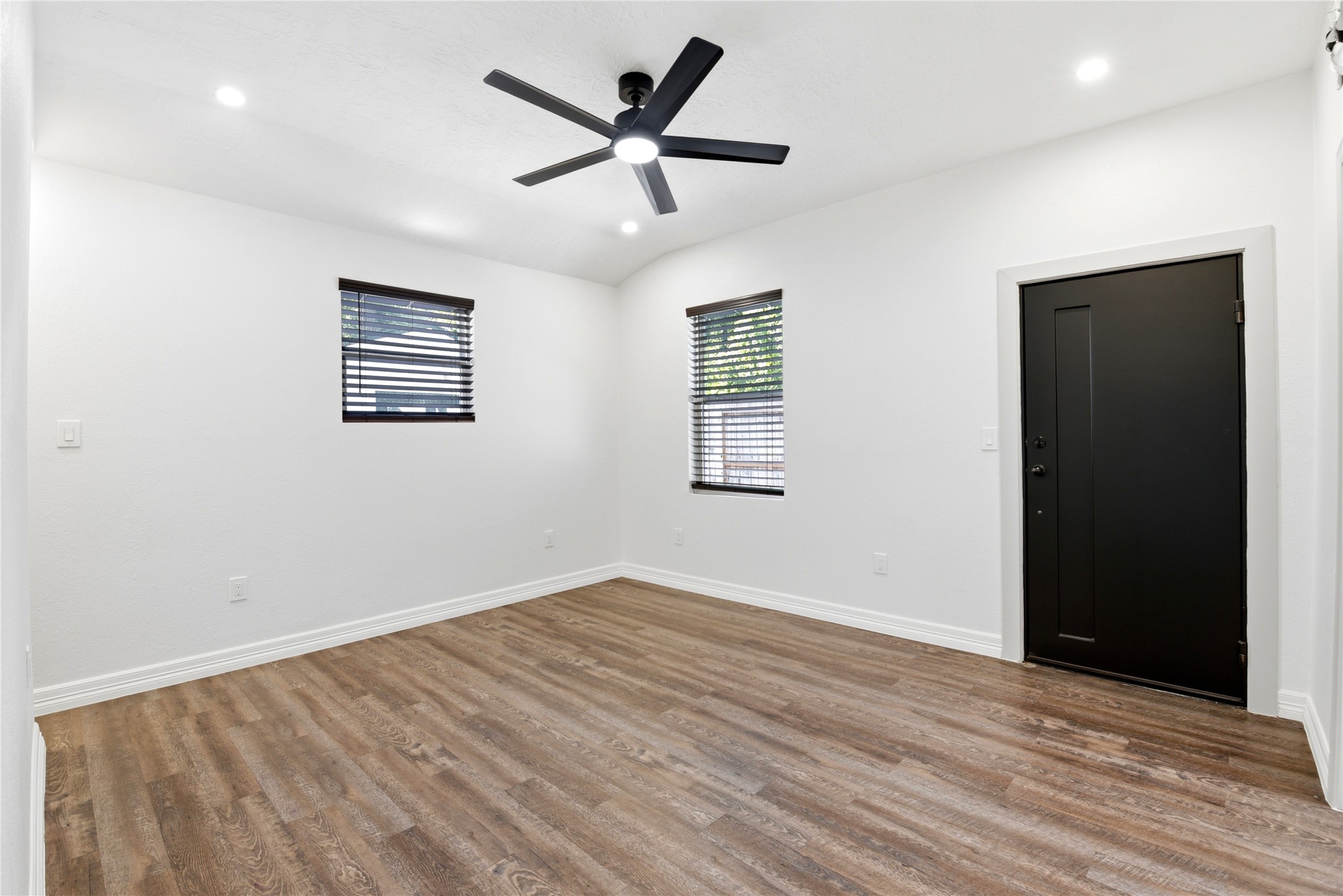 9403 Spaulding Street Houston, TX 77016 - Photo 32 of 50 This room features a modern, minimalist design with white walls, wood flooring, and a black ceiling fan. Two windows with blinds provide natural light, and there's a sleek black door, creating a contemporary feel.