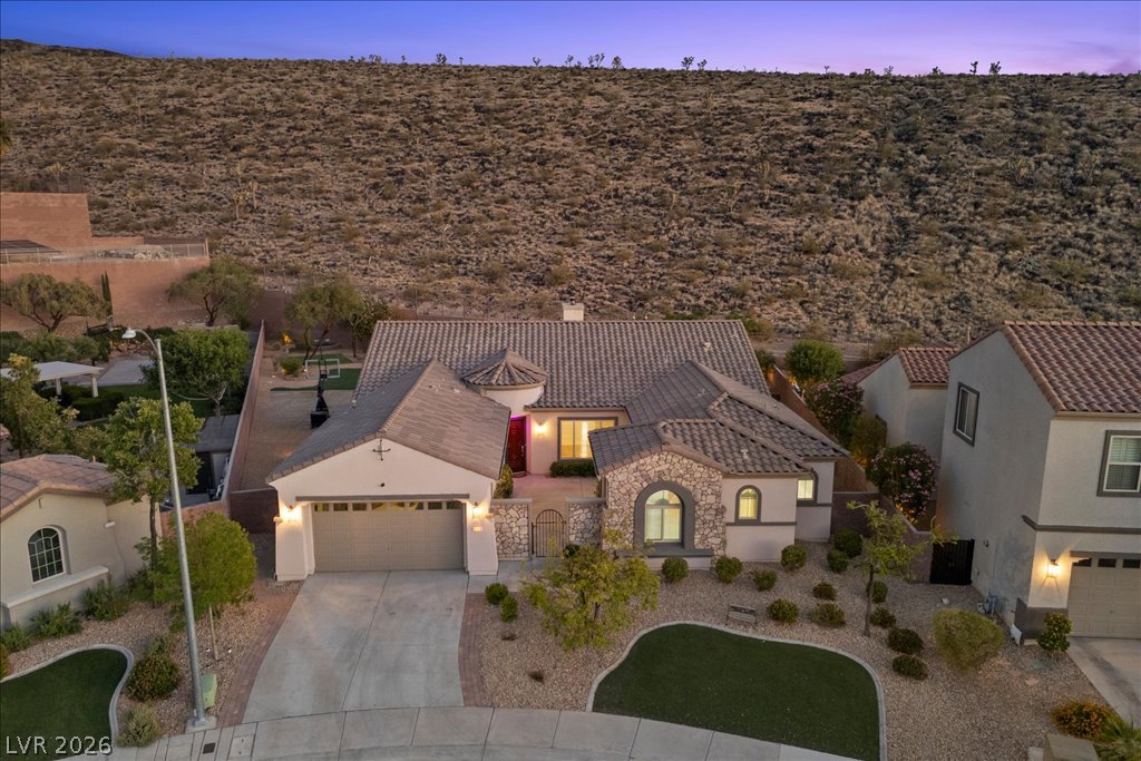 2125 De Rouge Avenue Henderson, NV 89044 - Photo 86 of 96 A desert-modern residence with a stone facade, tile roof, and a meticulously manicured front yard offers captivating views of the rugged hillside.
