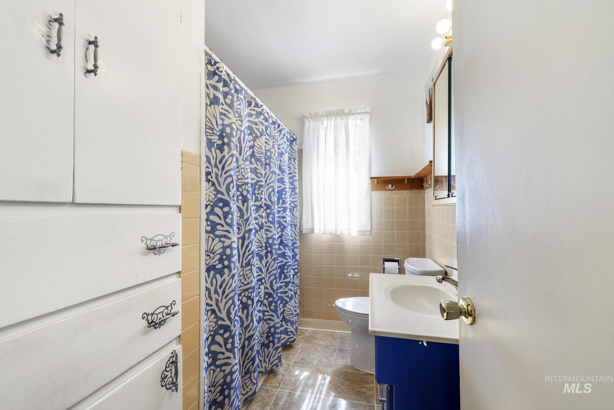 19591 Queen Crown Road Carey, ID 83320 - Photo 14 of 37 Bathroom featuring a white built-in storage cabinet, beige wall tiles, a window with sheer curtains, a vanity with a white sink, and a mirrored medicine cabinet