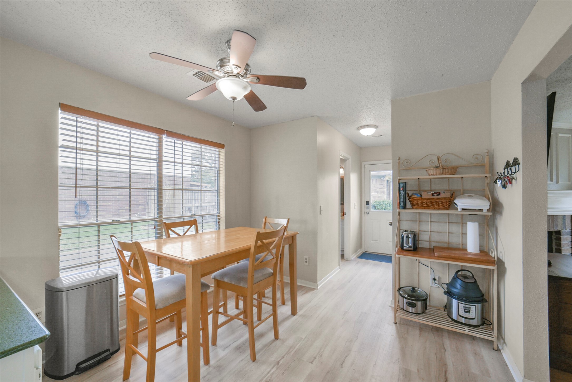 10410 Dude Road Houston, TX 77064 - Photo 13 of 45 Bright dining area with a ceiling fan, featuring a wooden table and chairs. Large windows let in natural light, and there's a view of the backyard. The space includes a utility shelf and has easy access to the back door. Light wood flooring adds warmth.