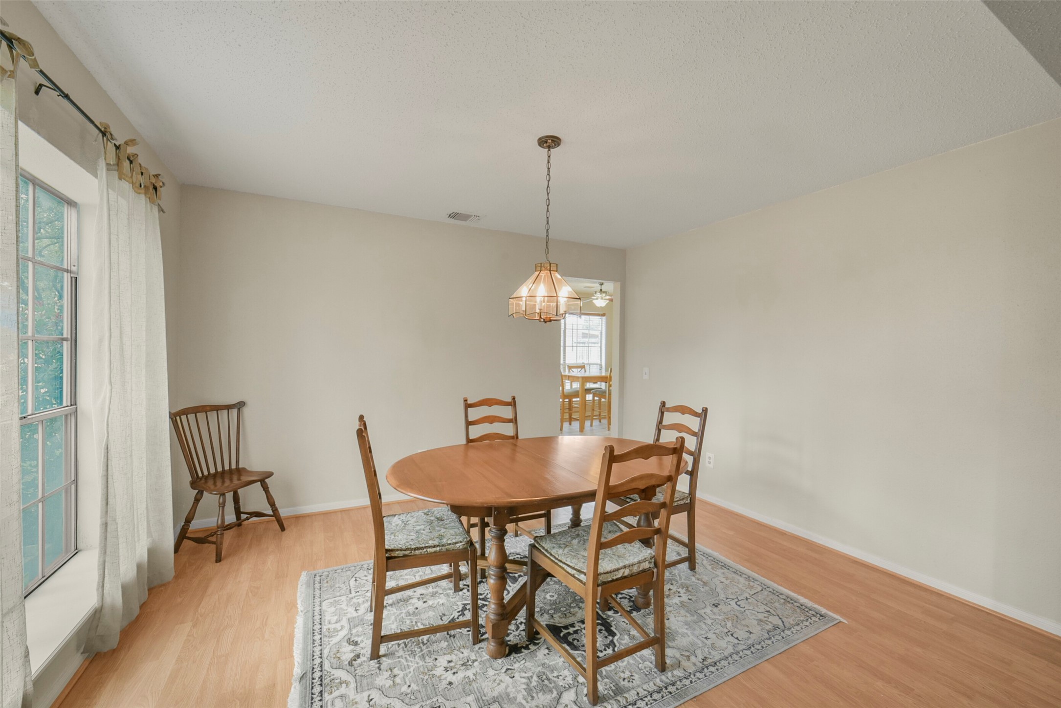 10410 Dude Road Houston, TX 77064 - Photo 16 of 45 This dining room features a light wood floor, a central round table with four chairs, and a large window with natural light. It has neutral walls and a classic hanging light fixture, creating a cozy, inviting space.