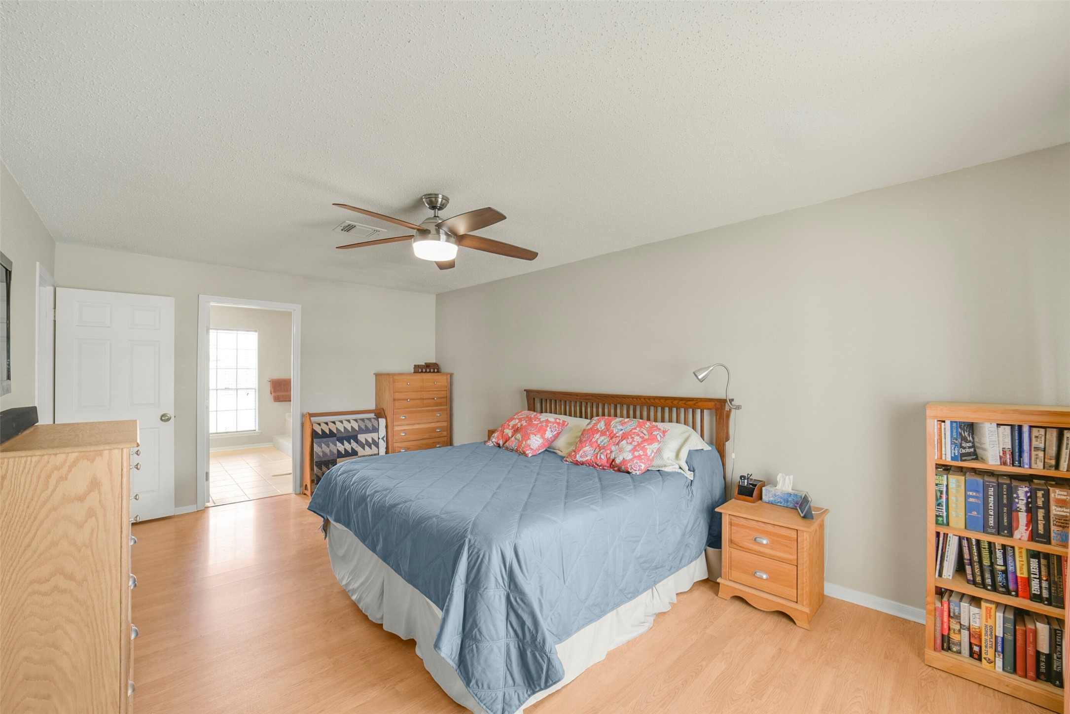 10410 Dude Road Houston, TX 77064 - Photo 19 of 45 This bedroom features a spacious layout with light wood flooring and neutral walls. It includes a ceiling fan, ample natural light, and wooden furniture, including dressers and a bookcase. A doorway leads to an adjacent room with large windows.